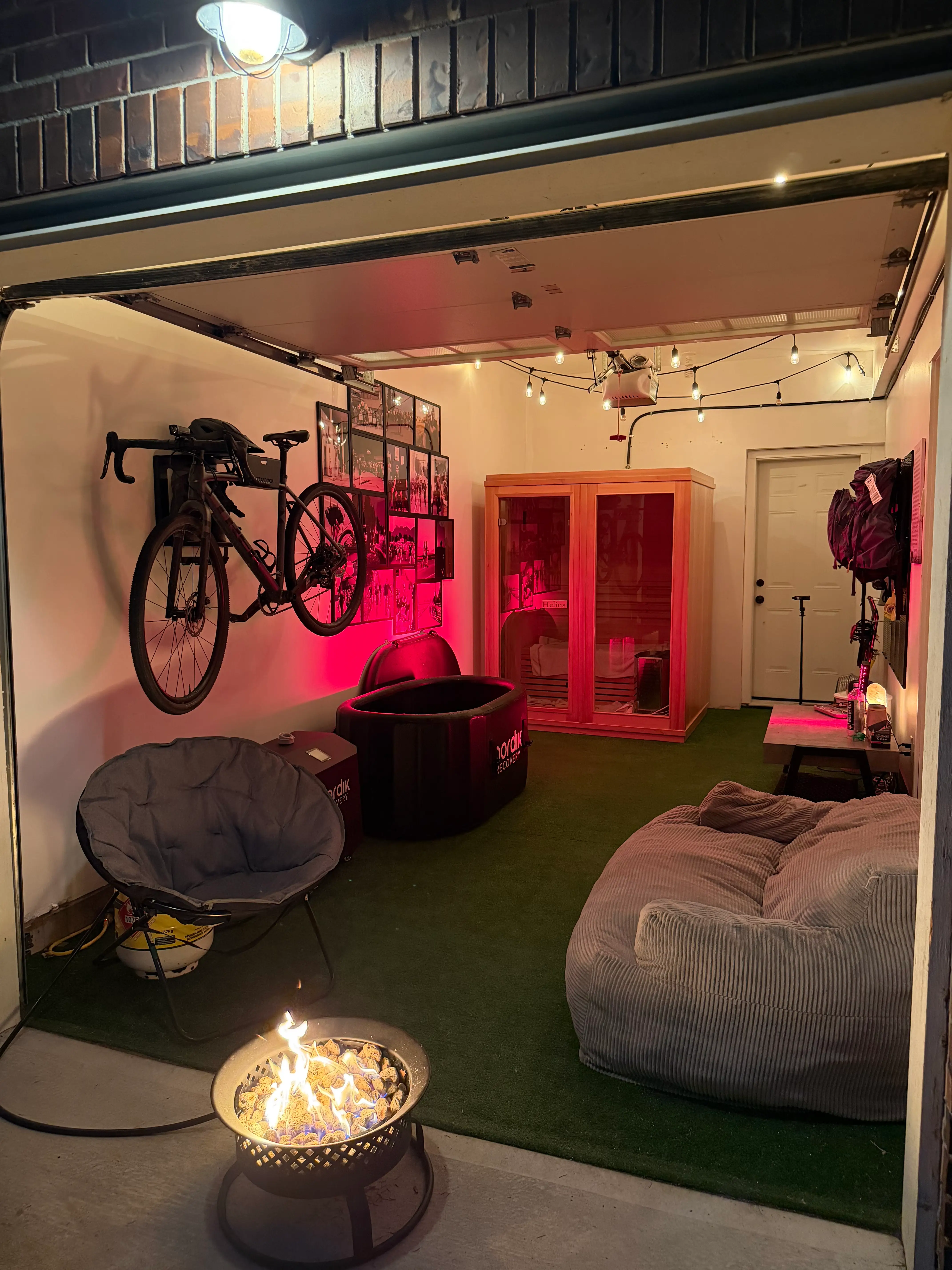 An open garage lounge with wall-mounted bicycle, fire pit, seating, sauna, string lights, and artificial turf.