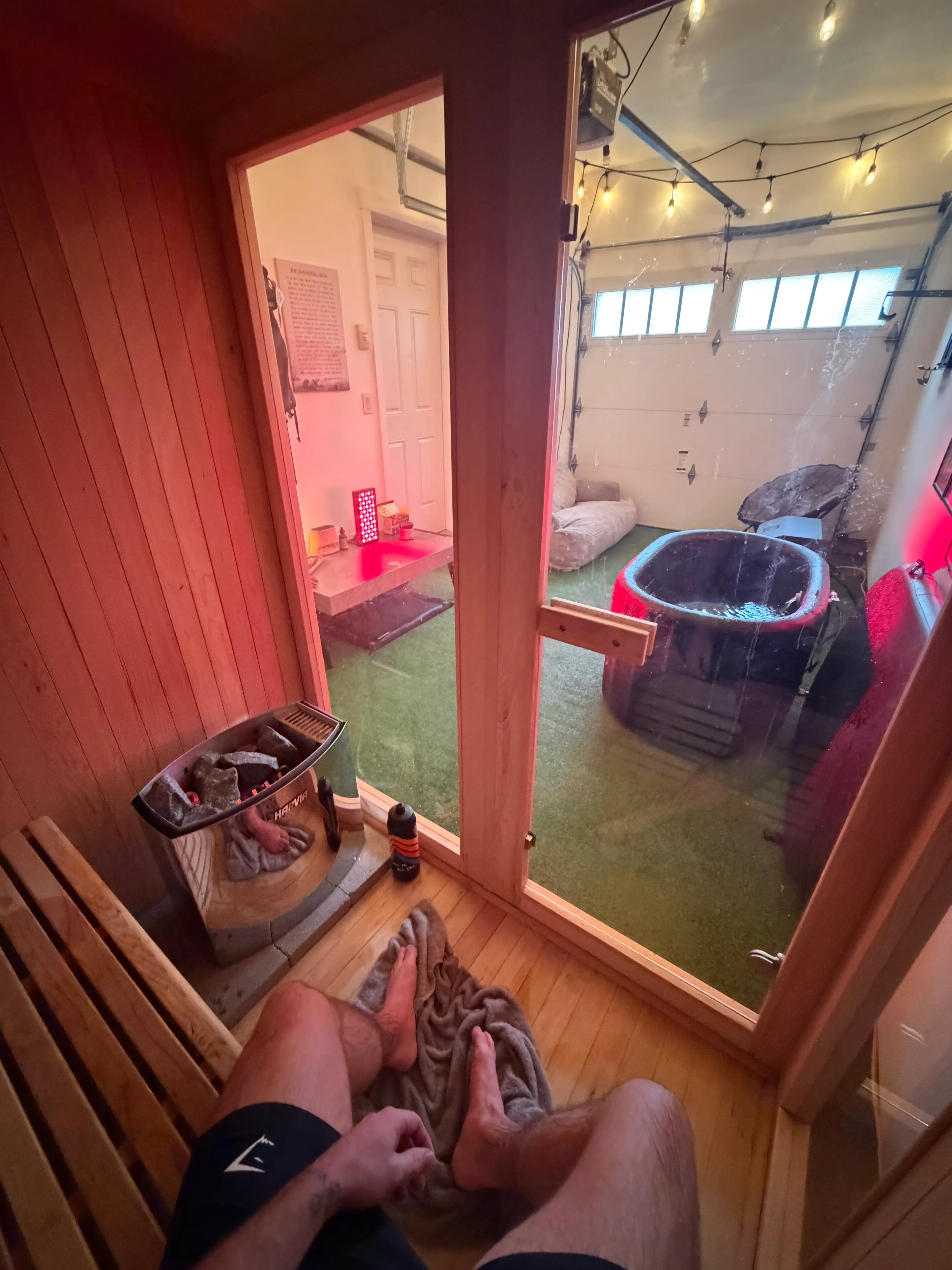 Sauna interior overlooks a garage spa area with a black tub, string lights, and artificial turf.