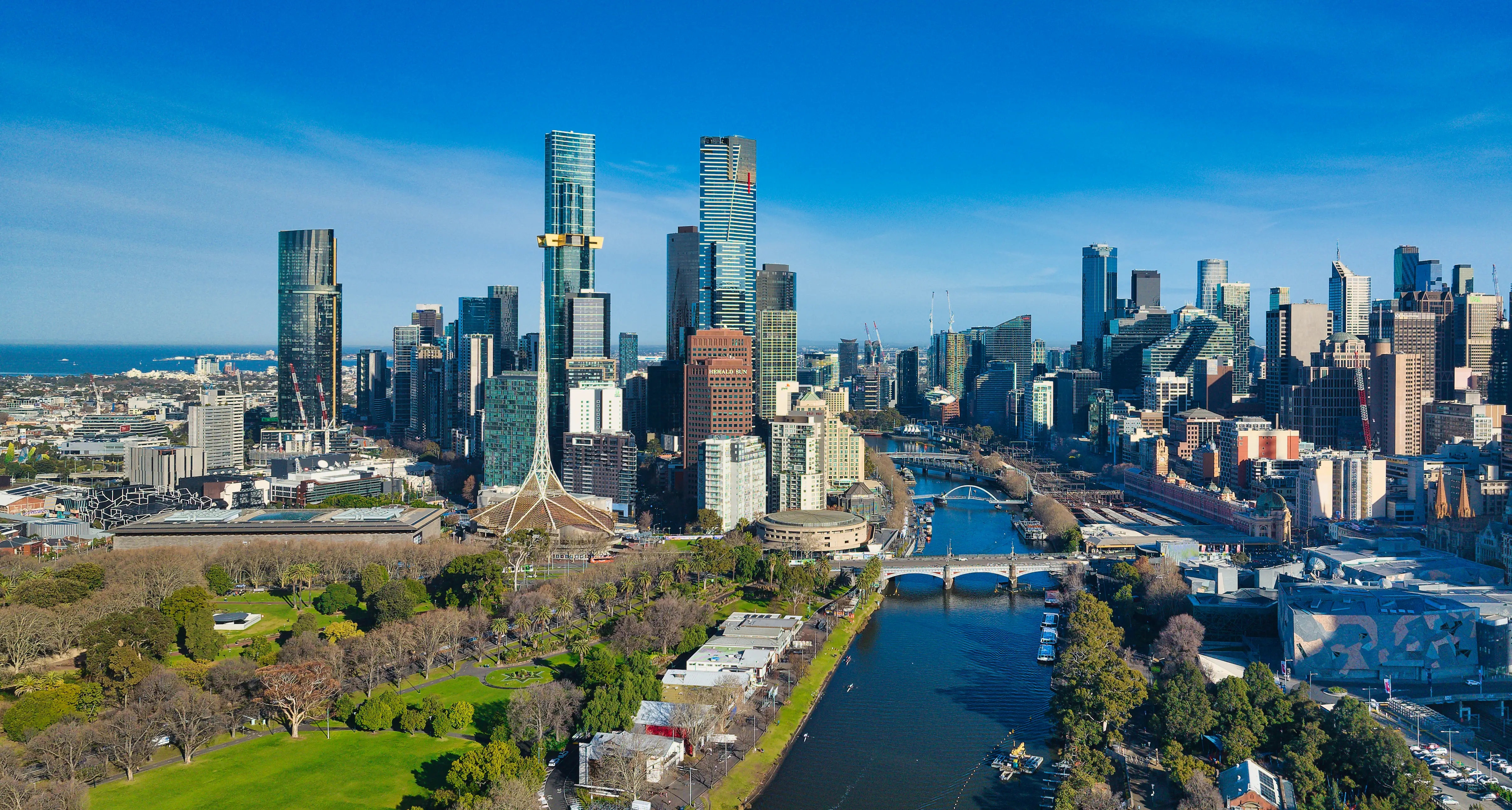Melbourne skyline rises beside the Yarra River with parkland, bridges, and the Arts Centre Melbourne spire in view.
