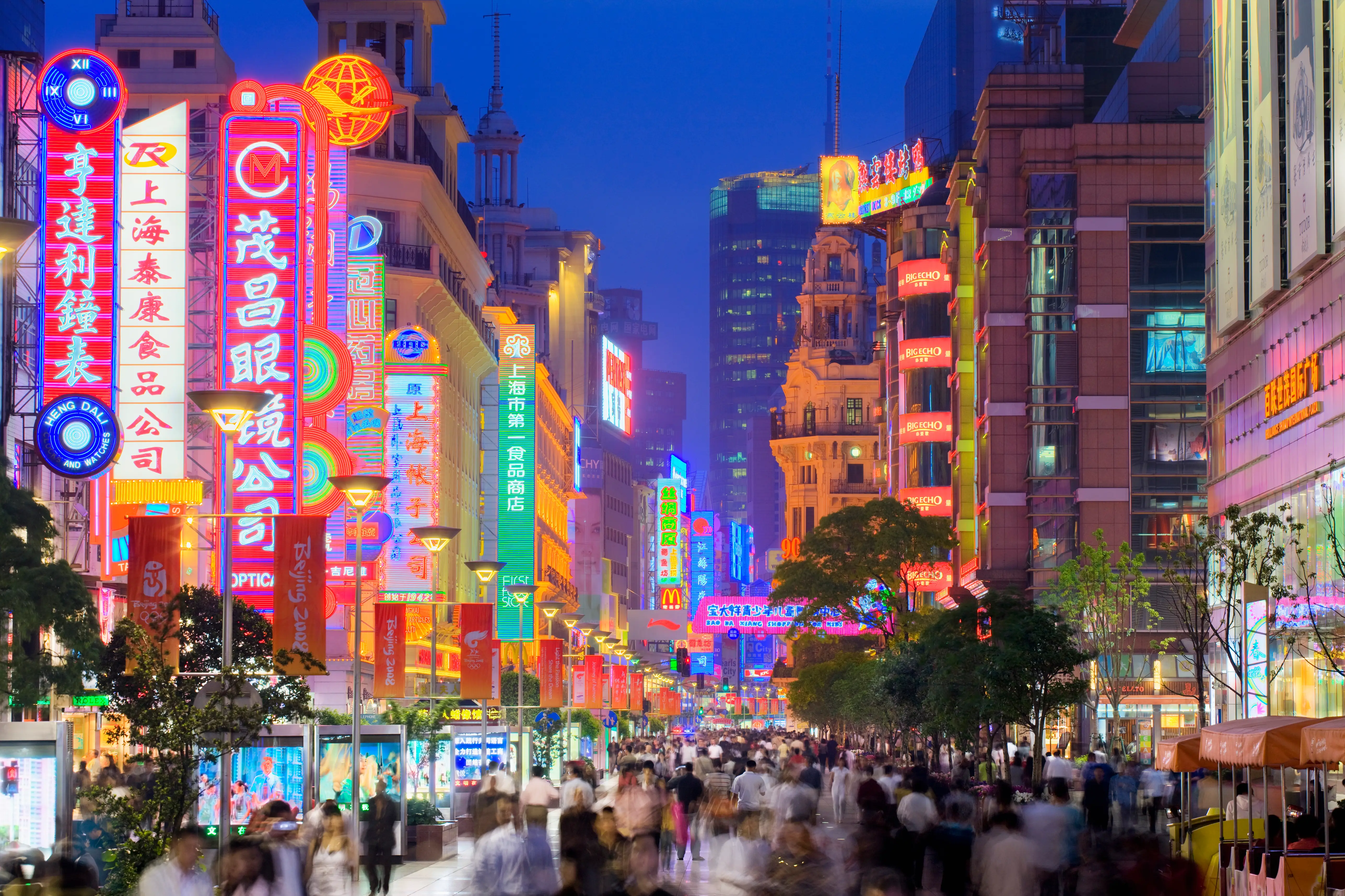 Neon Chinese storefront signs illuminate a crowded city shopping street at dusk with blurred pedestrians and tall buildings.