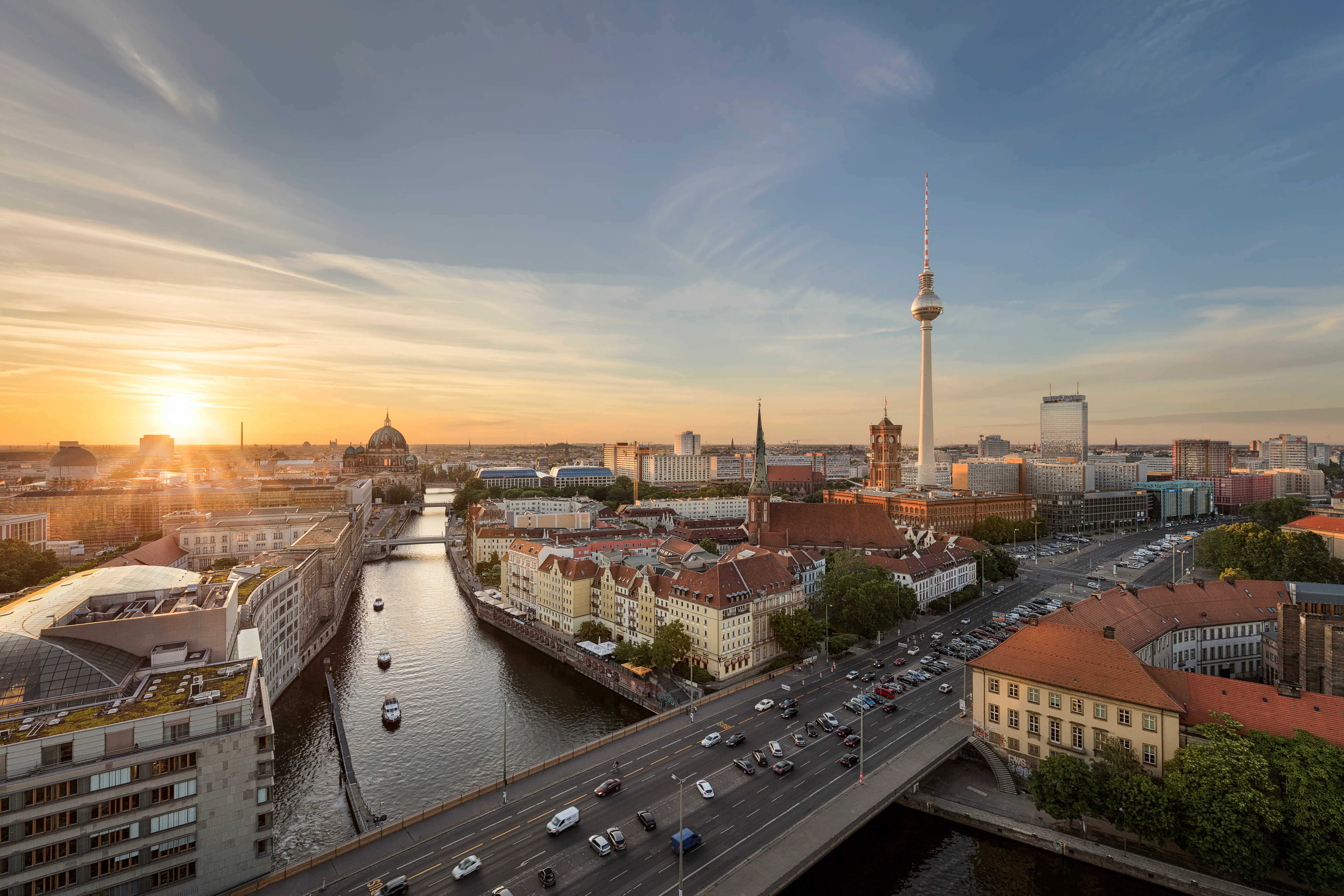 Berlin skyline at sunset with the Fernsehturm, Berliner Dom, Spree River, bridges, traffic, and red-roofed buildings.