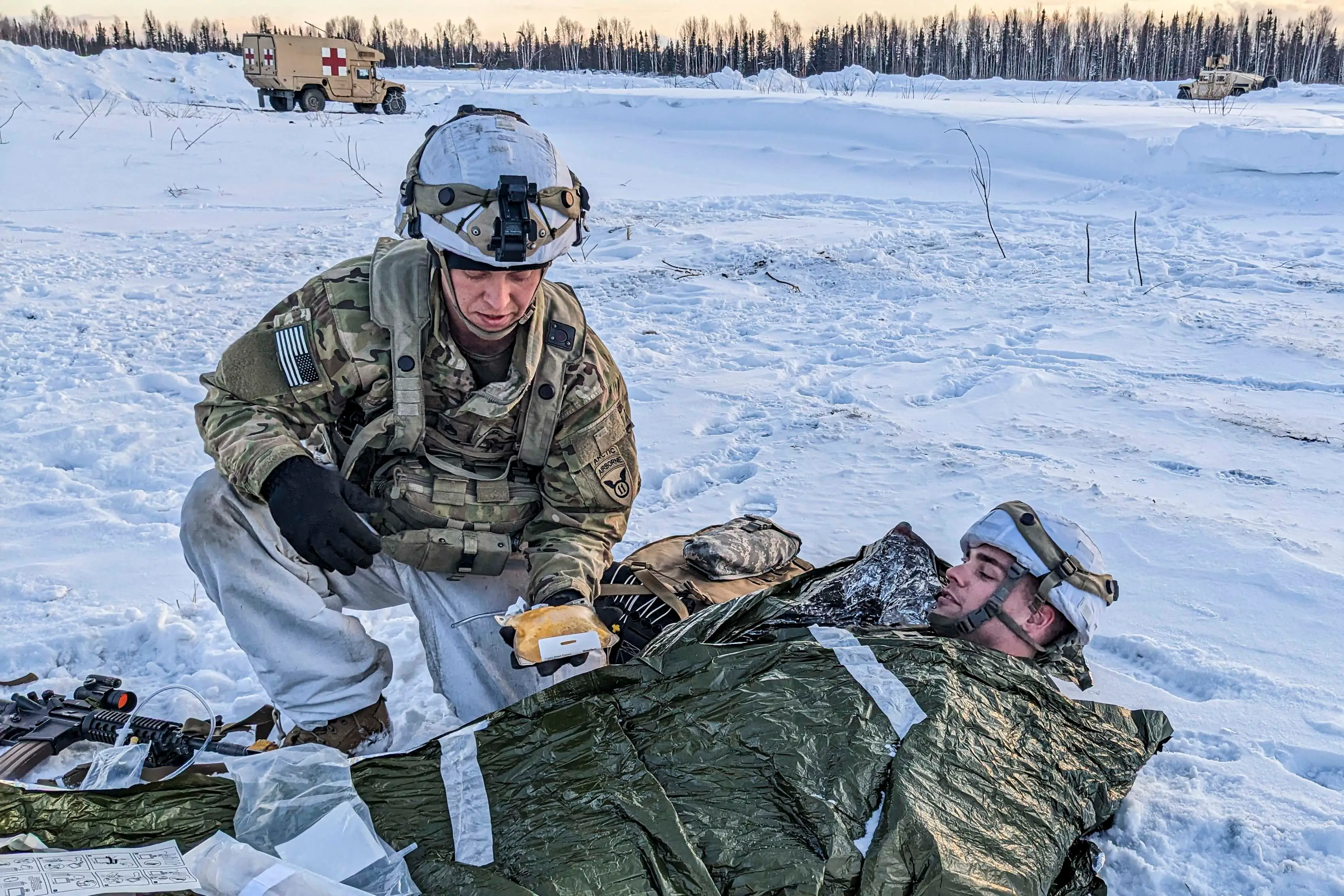 Service members provide snowy field medical aid to a person wrapped in an emergency blanket near marked medical vehicles.