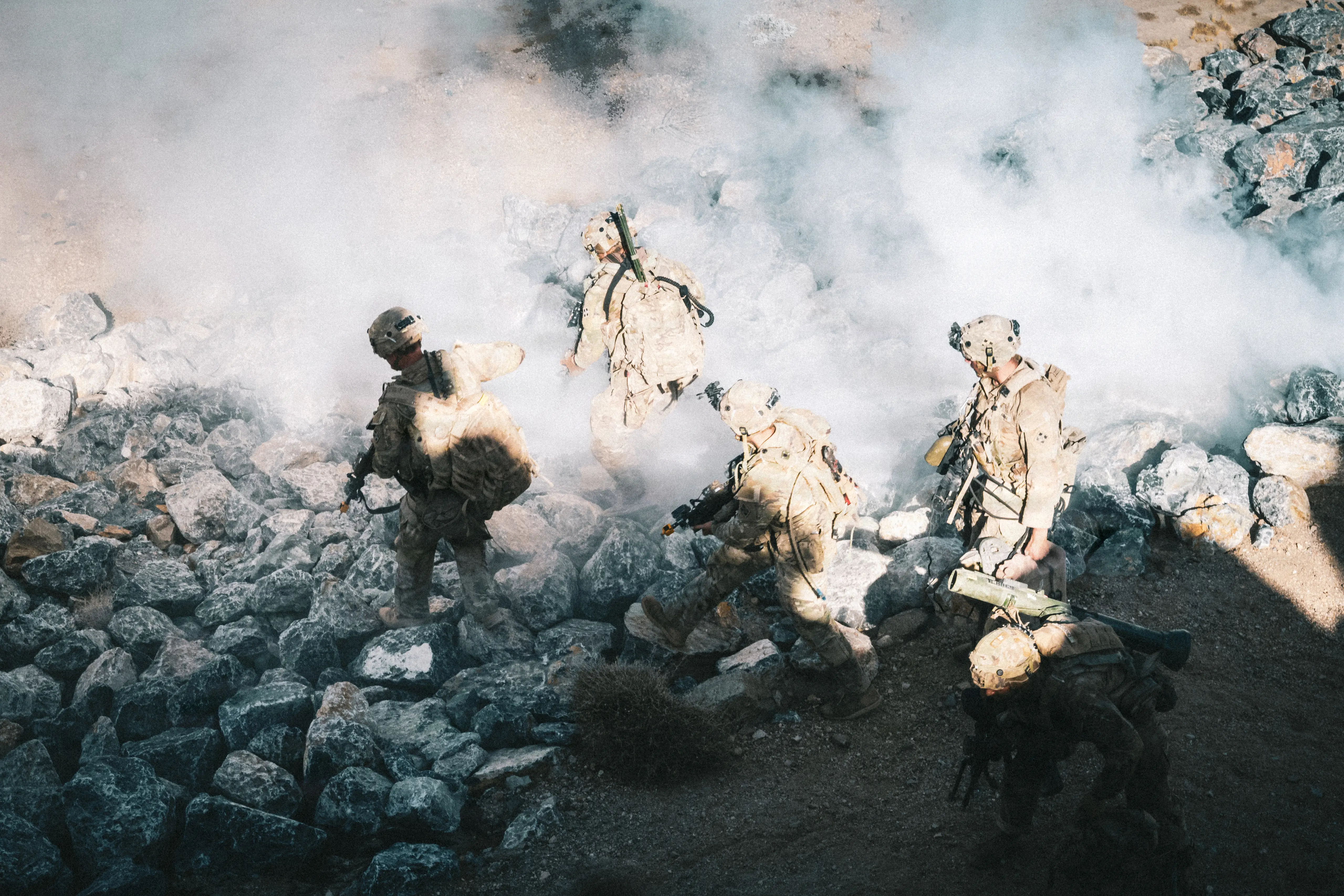 Four US Army soldiers walk down a rocky hill with smoke surrounding them.