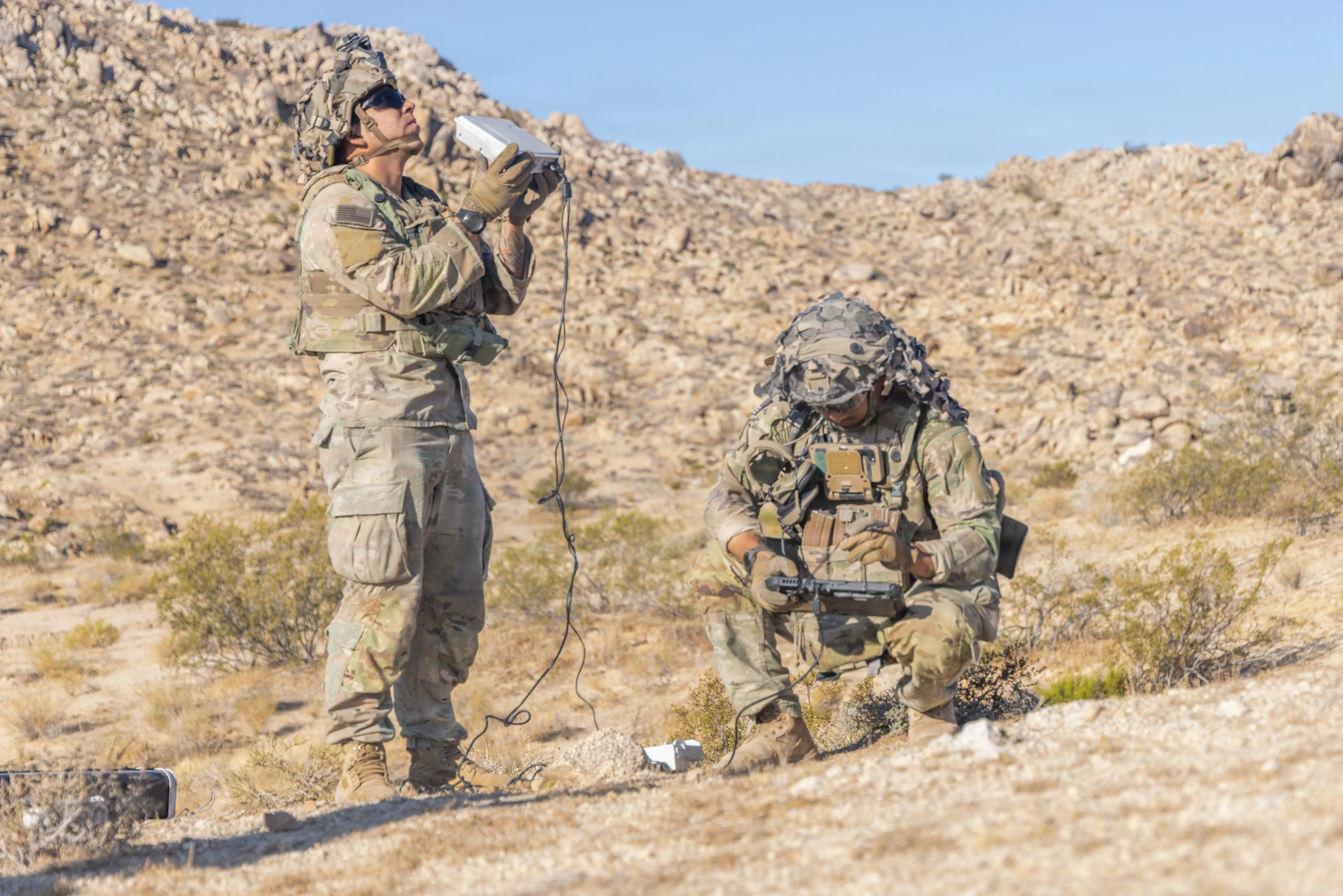 One US Army soldier stands holding a white box while another soldier crouches holding a drone controller. The soldiers are in the desert.