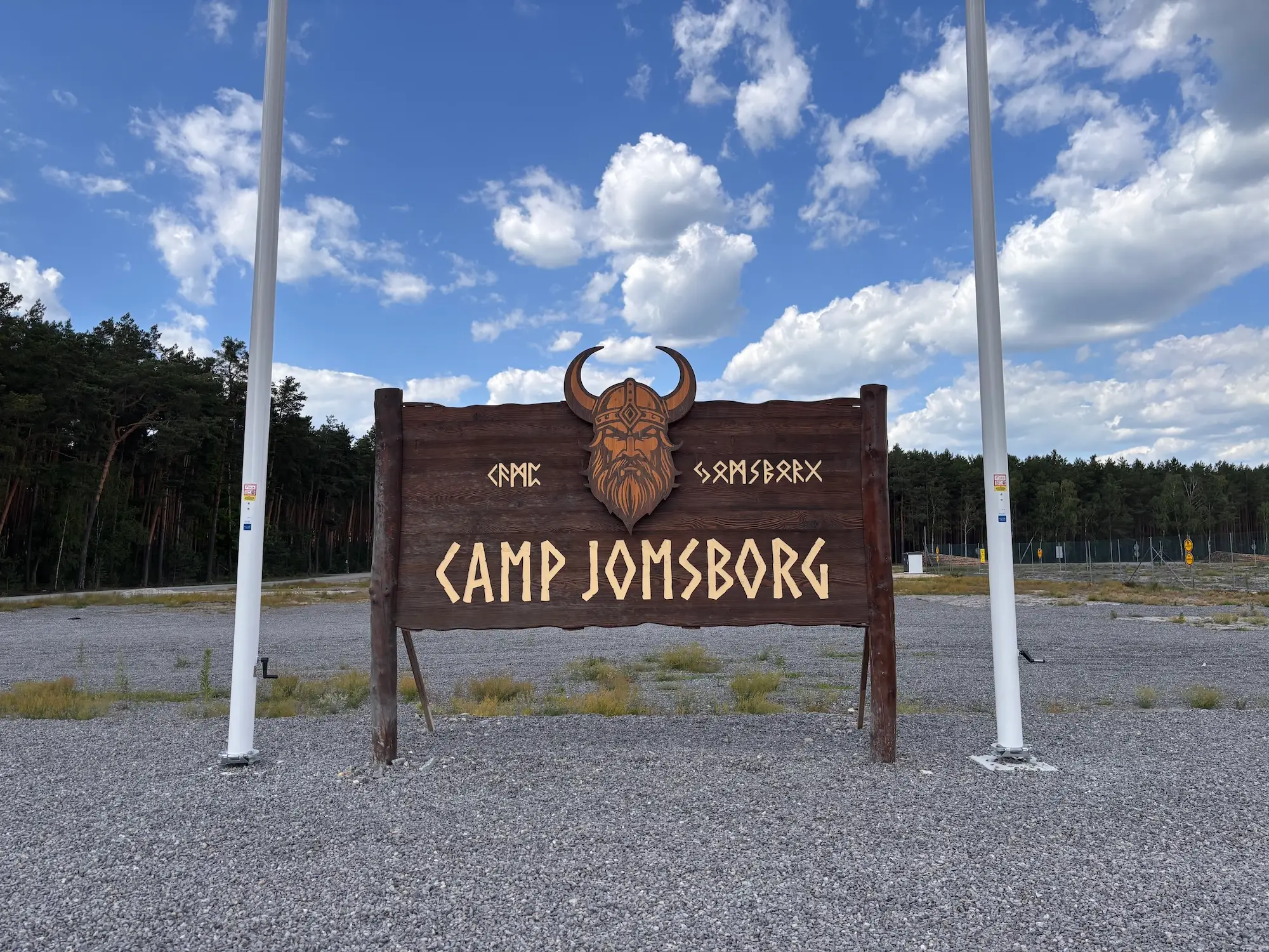 Wooden CAMP JOMSBORG entrance sign with a Viking head emblem stands between two flagpoles near a forest.