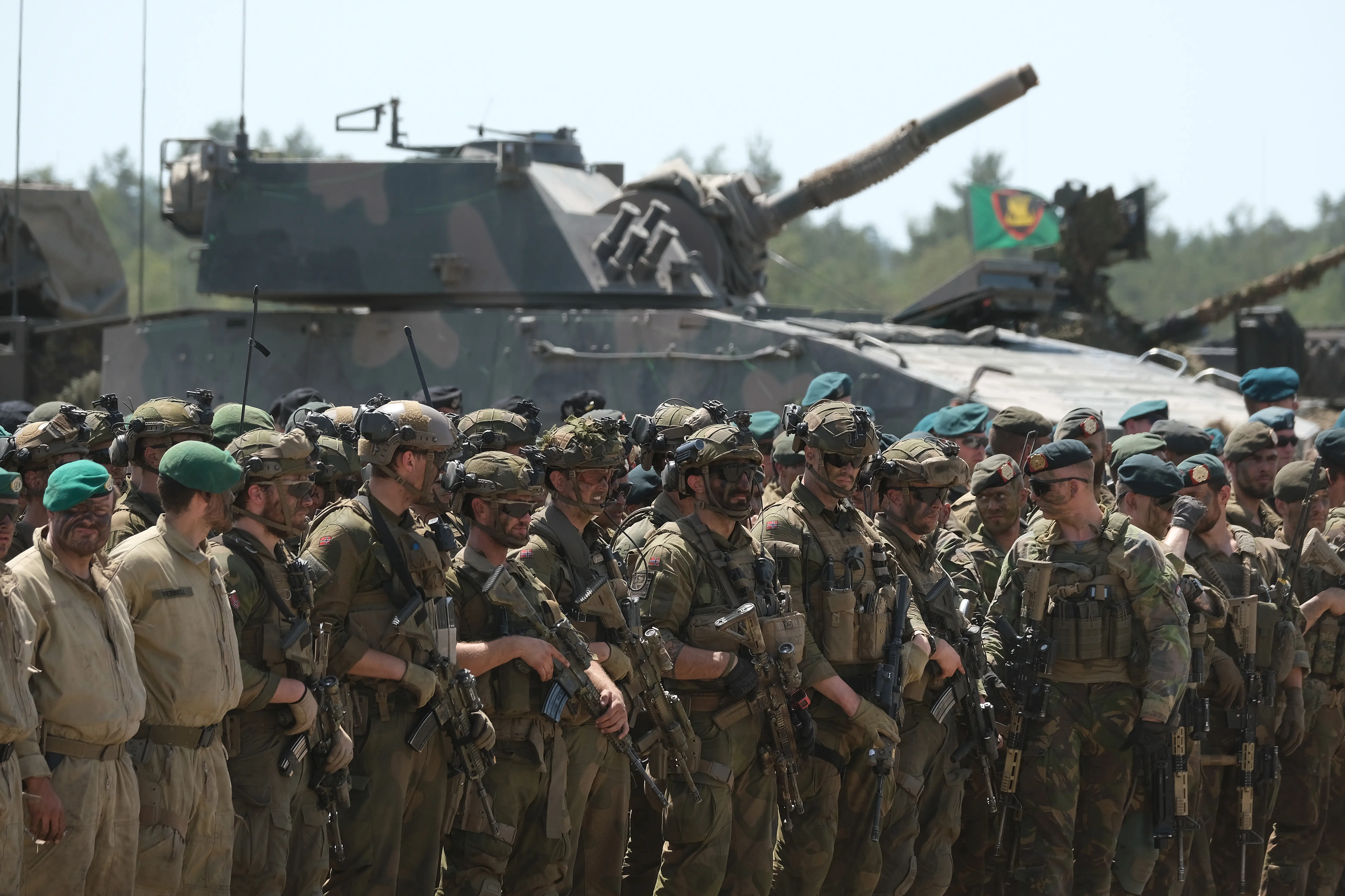 Uniformed soldiers carrying rifles stand in formation in front of armored vehicles and tank guns outdoors.