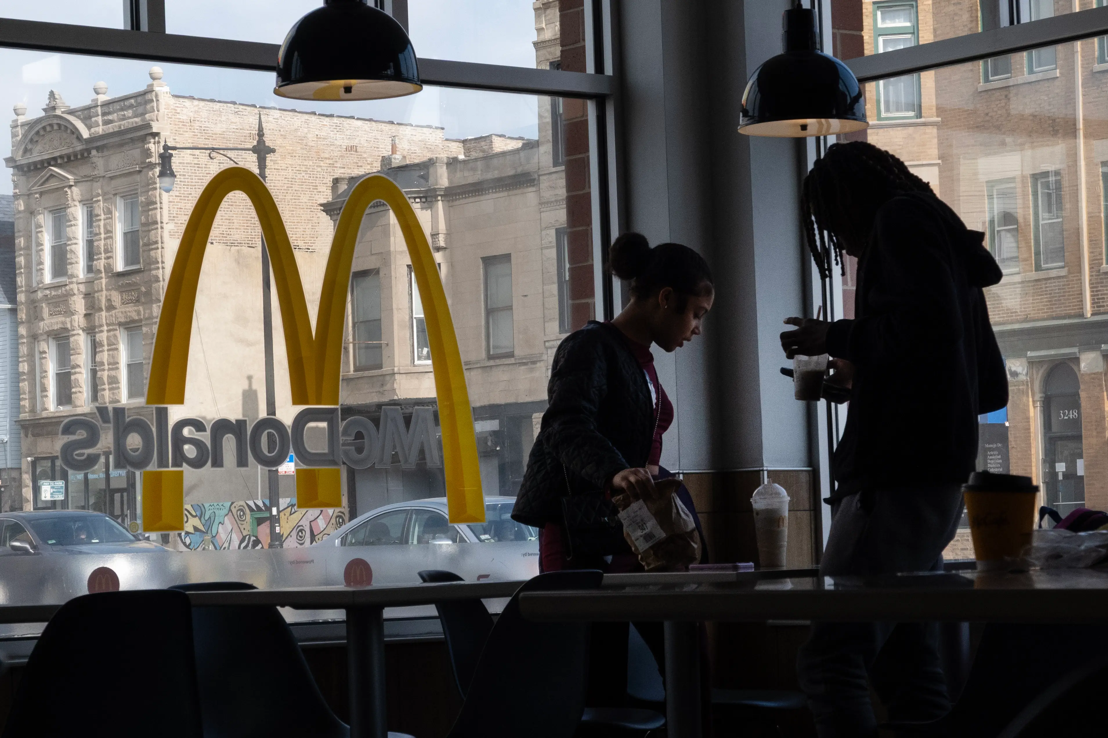 Two customers sit down inside of a McDonald's restaurant with the chain's logo visible in the window.