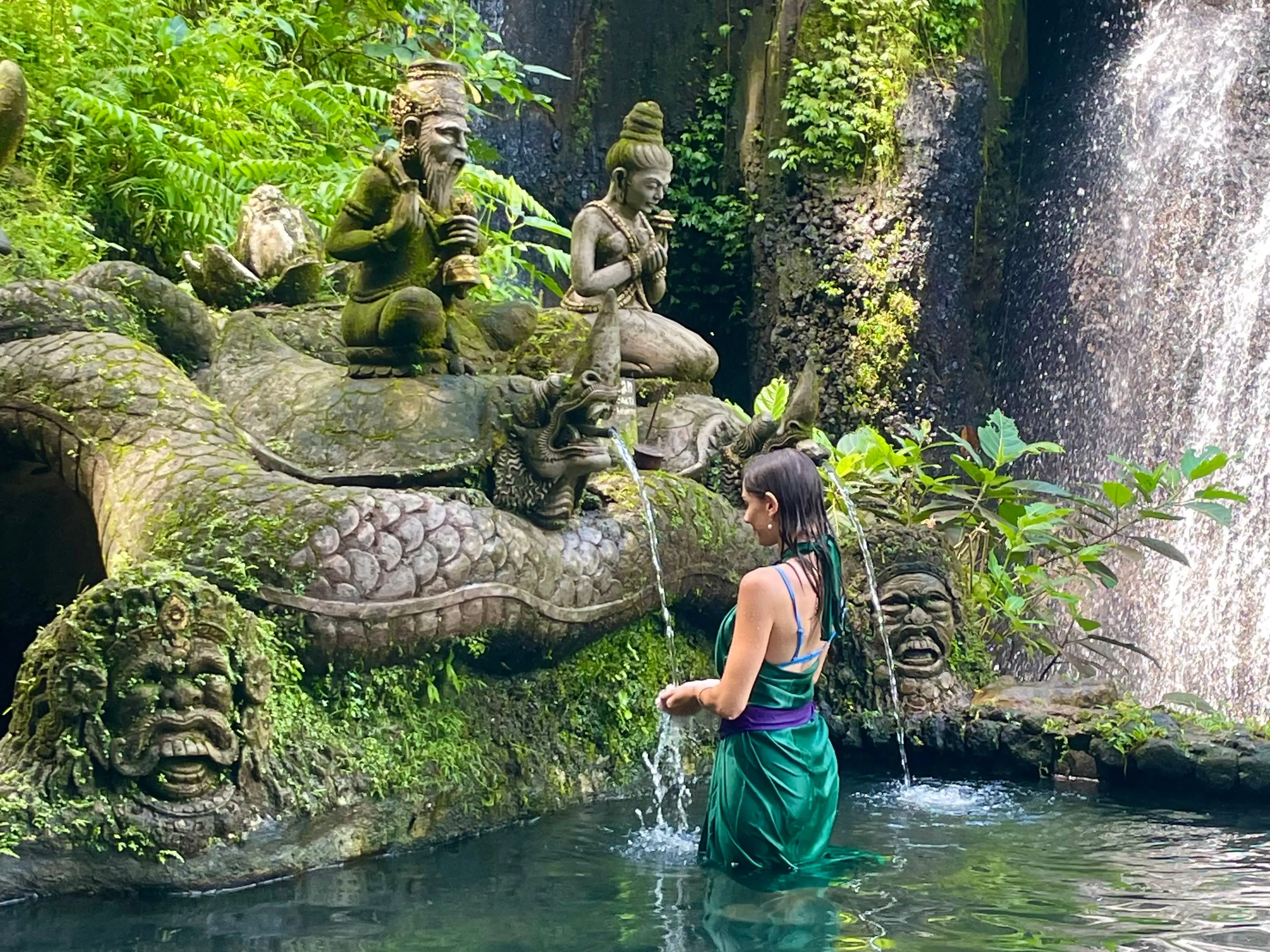 The writer standing in a pool in Bali, surrounded by moss and statues.