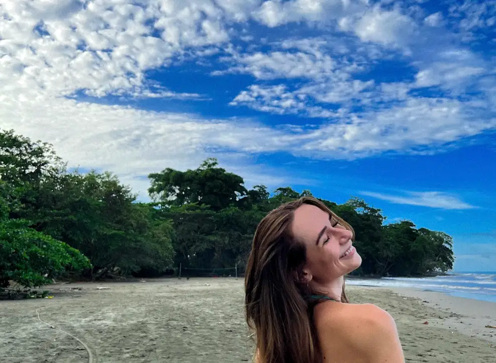 The writer stands on a sandy beach with trees, ocean water, and a cloud-filled blue sky.