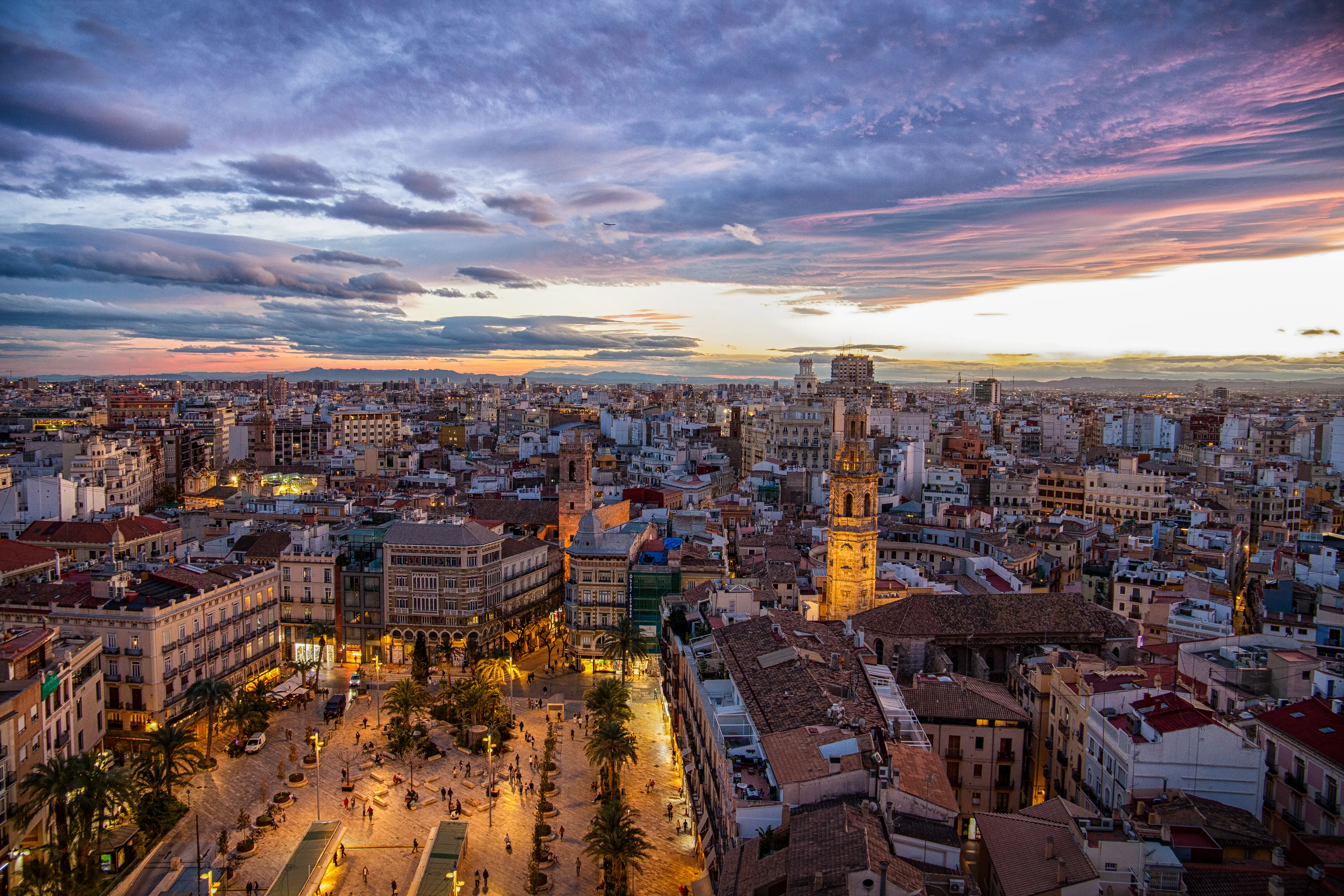 Valencia cityscape at sunset with the illuminated El Miguelete bell tower rising above dense urban streets.