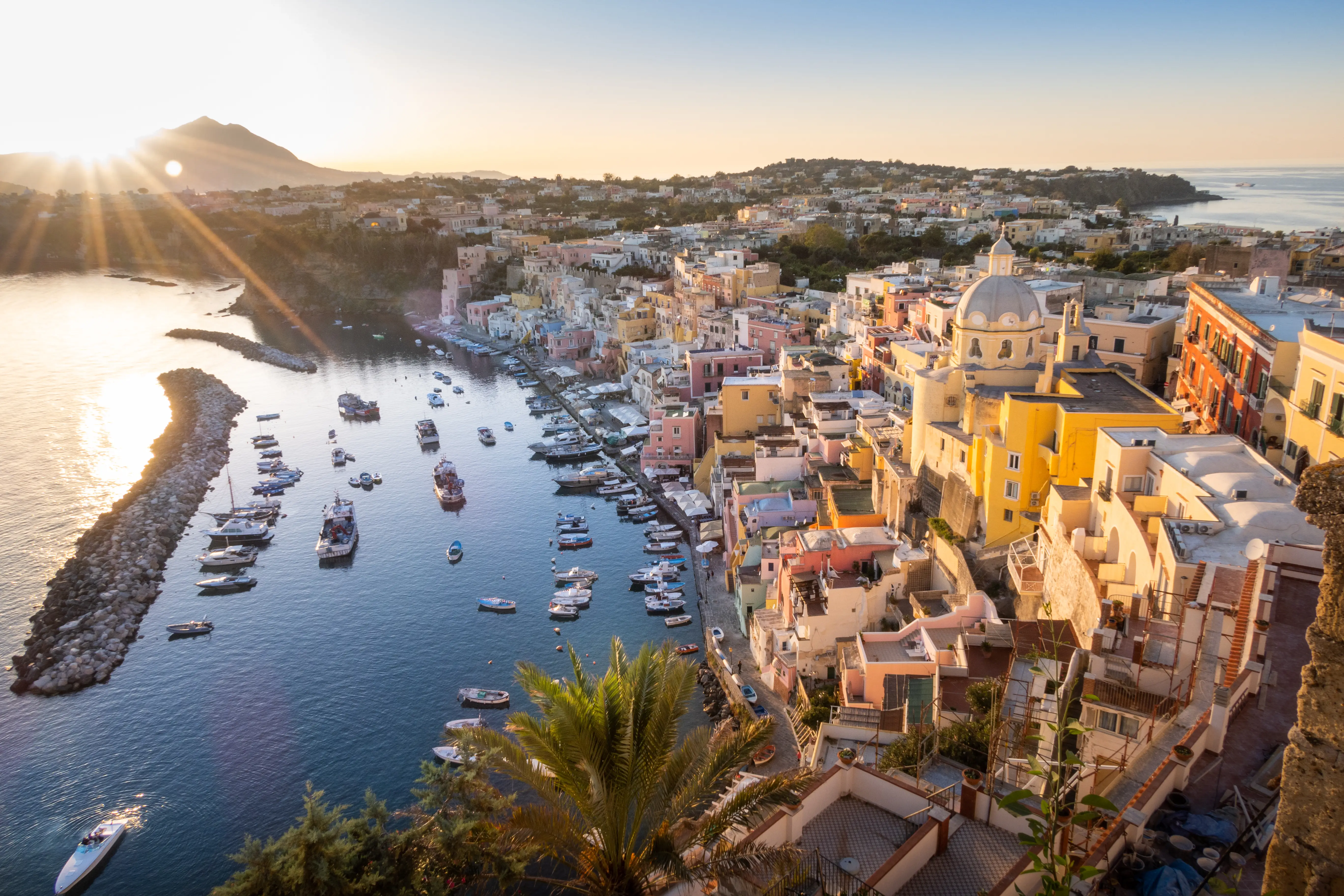 Sunlit harbor with boats and pastel hillside buildings surrounding a domed church on a coastal Mediterranean village.