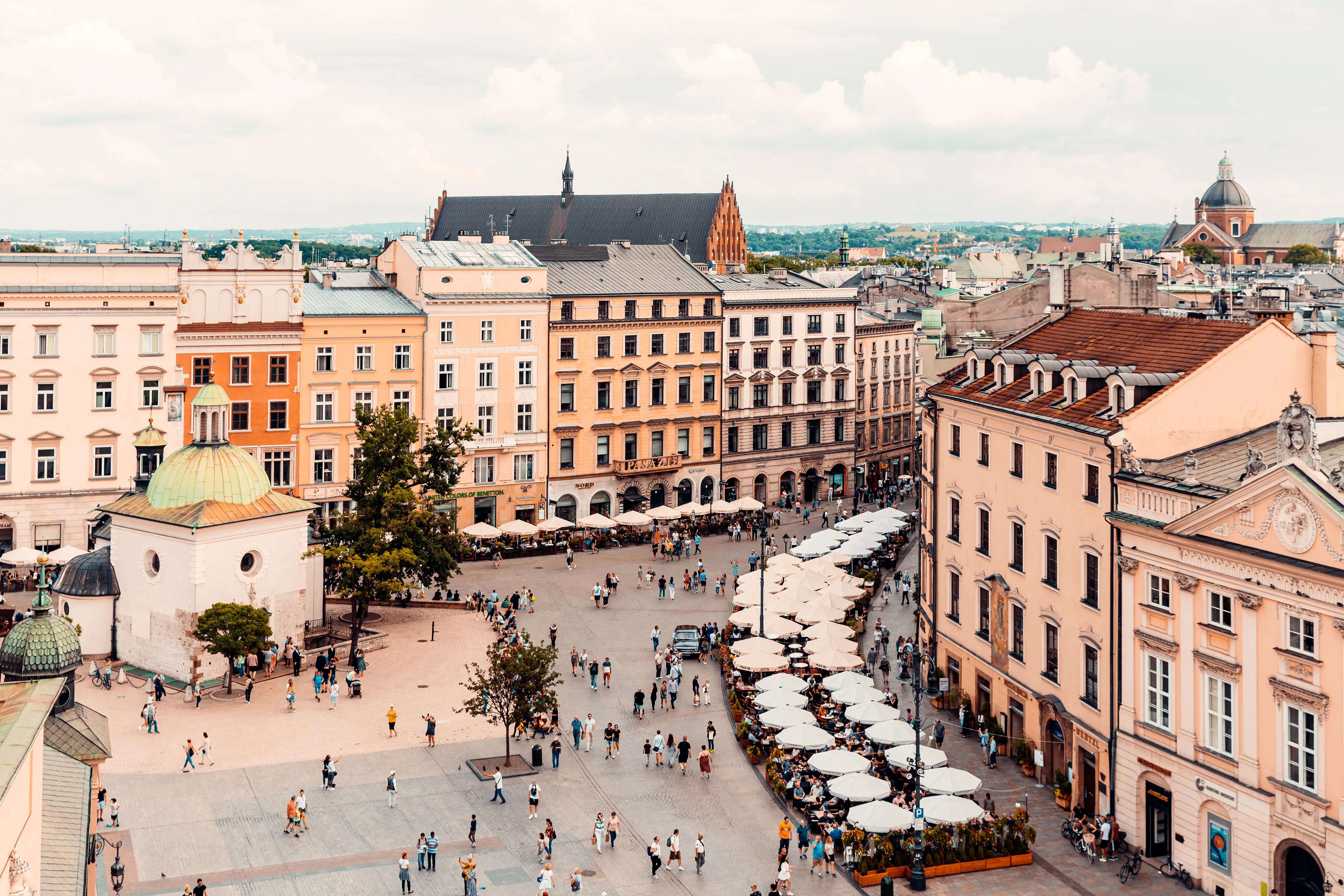 Kraków Main Market Square with historic buildings, church domes, café umbrellas, and pedestrians viewed from above.