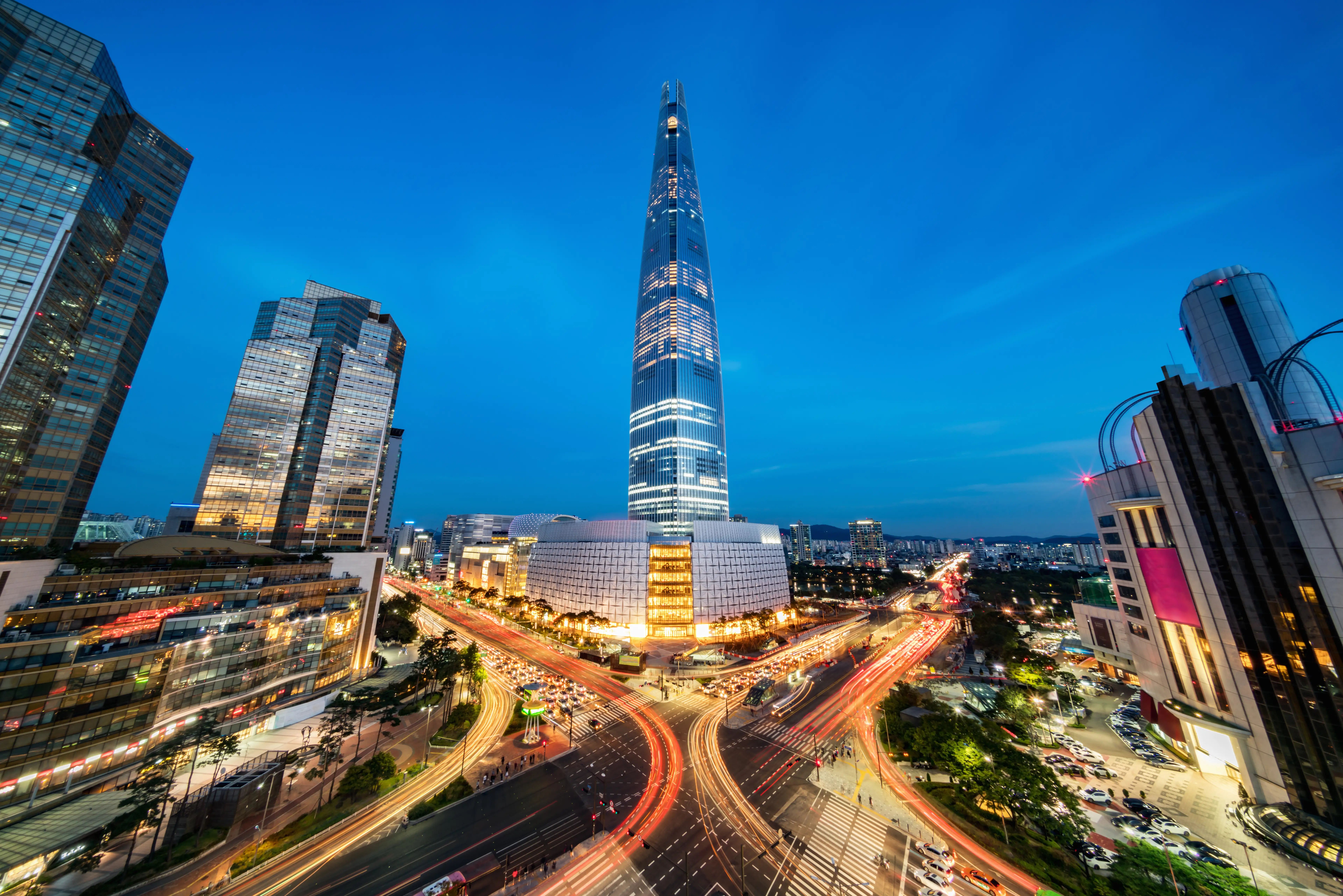 Lotte World Tower rises above illuminated Seoul streets with traffic light trails at dusk.