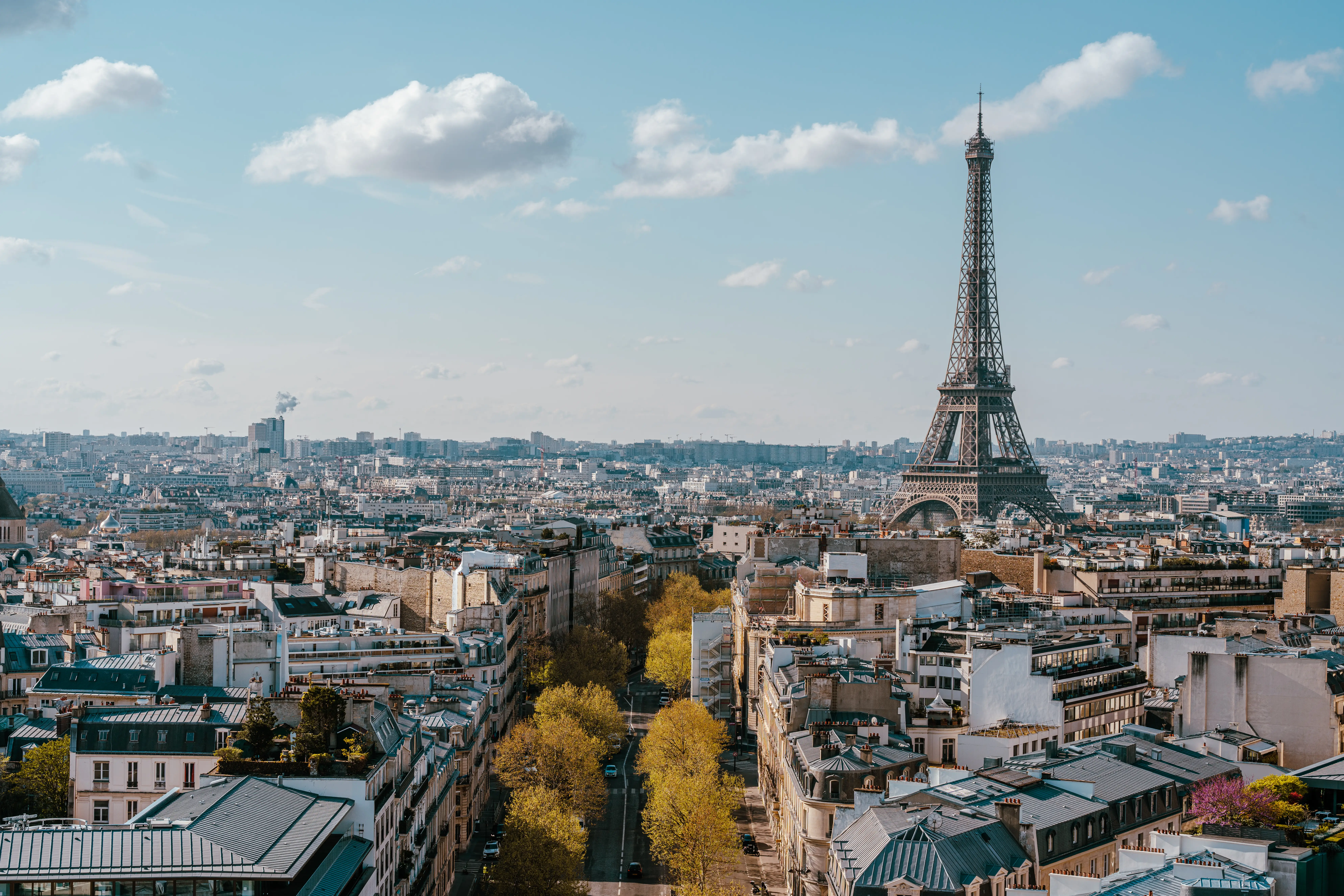 Paris cityscape with the Eiffel Tower rising above rooftops and a tree-lined avenue under a partly cloudy sky.