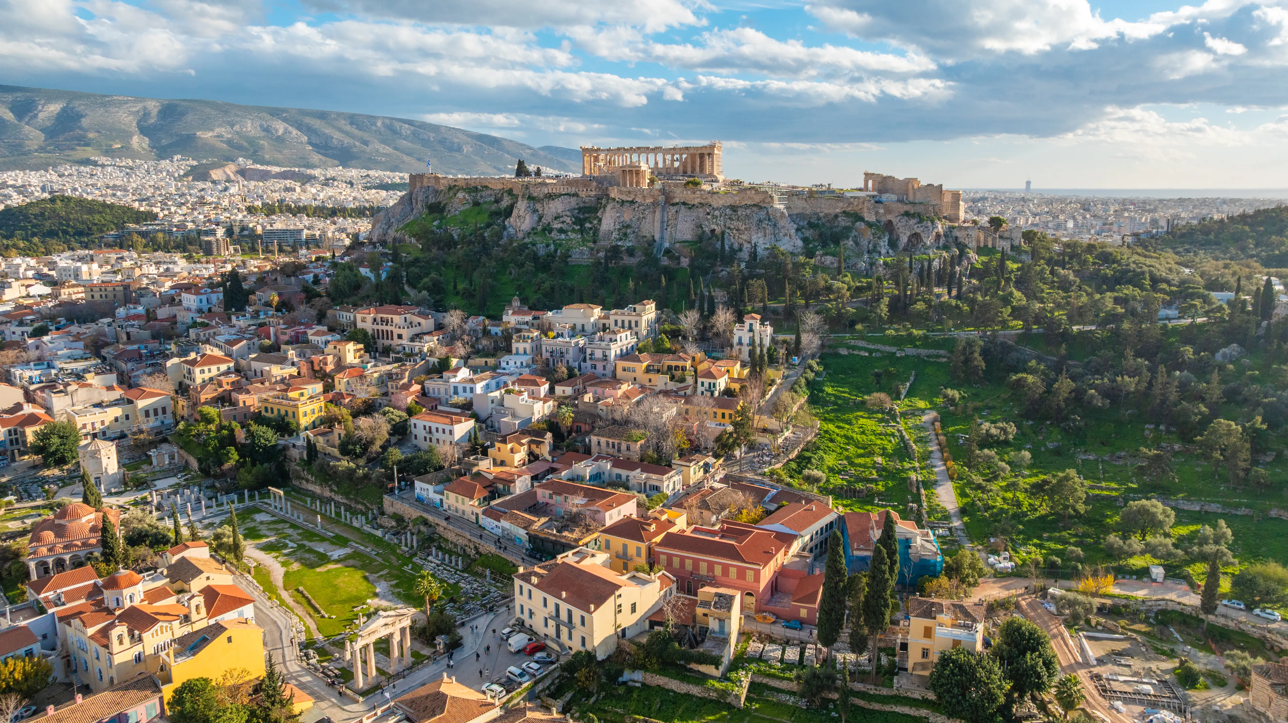 Athens cityscape with the Acropolis and Parthenon above historic neighborhoods under a partly cloudy sky.