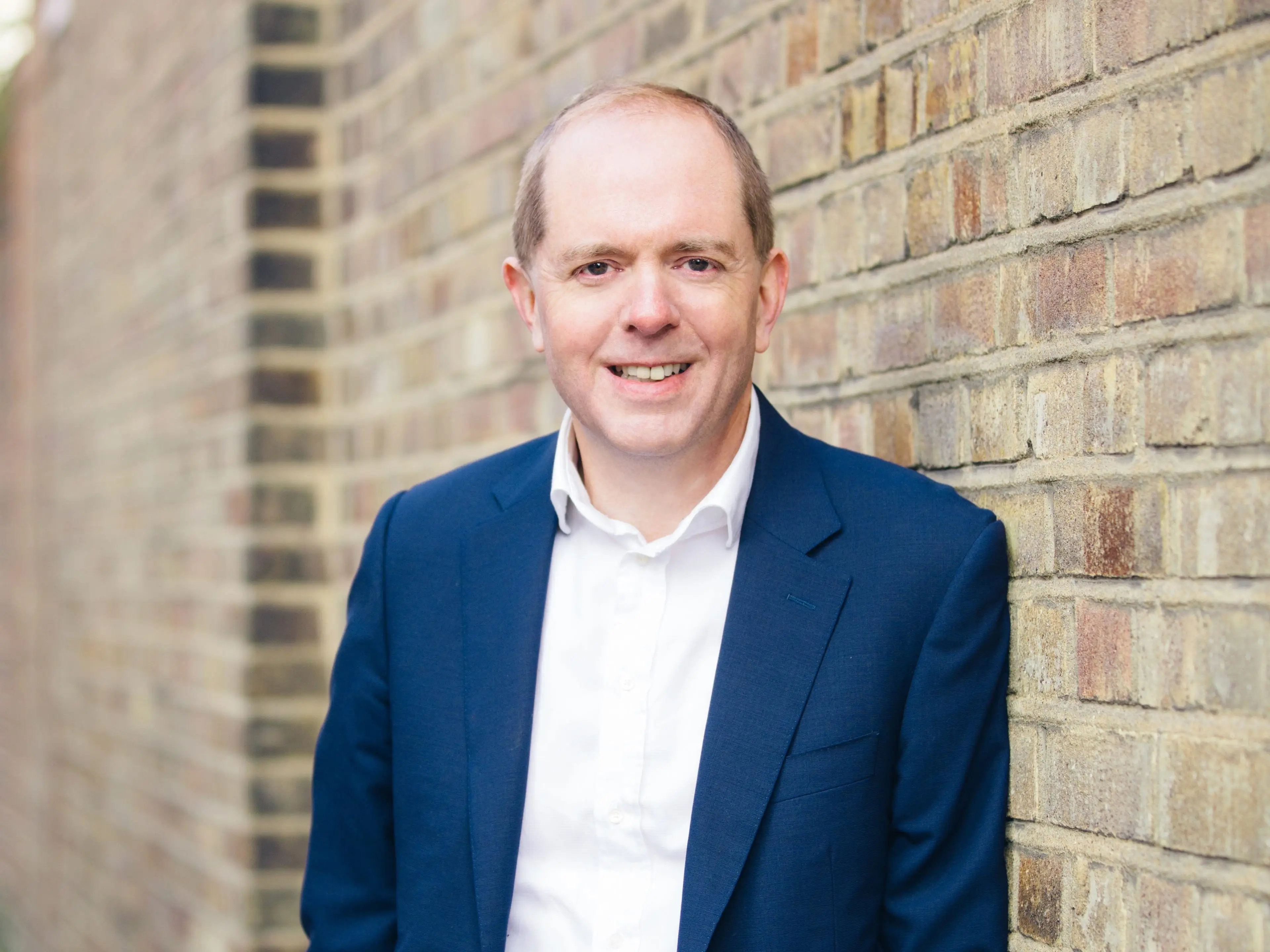 A person in a blue suit and white shirt stands leaning against a brick wall outdoors.
