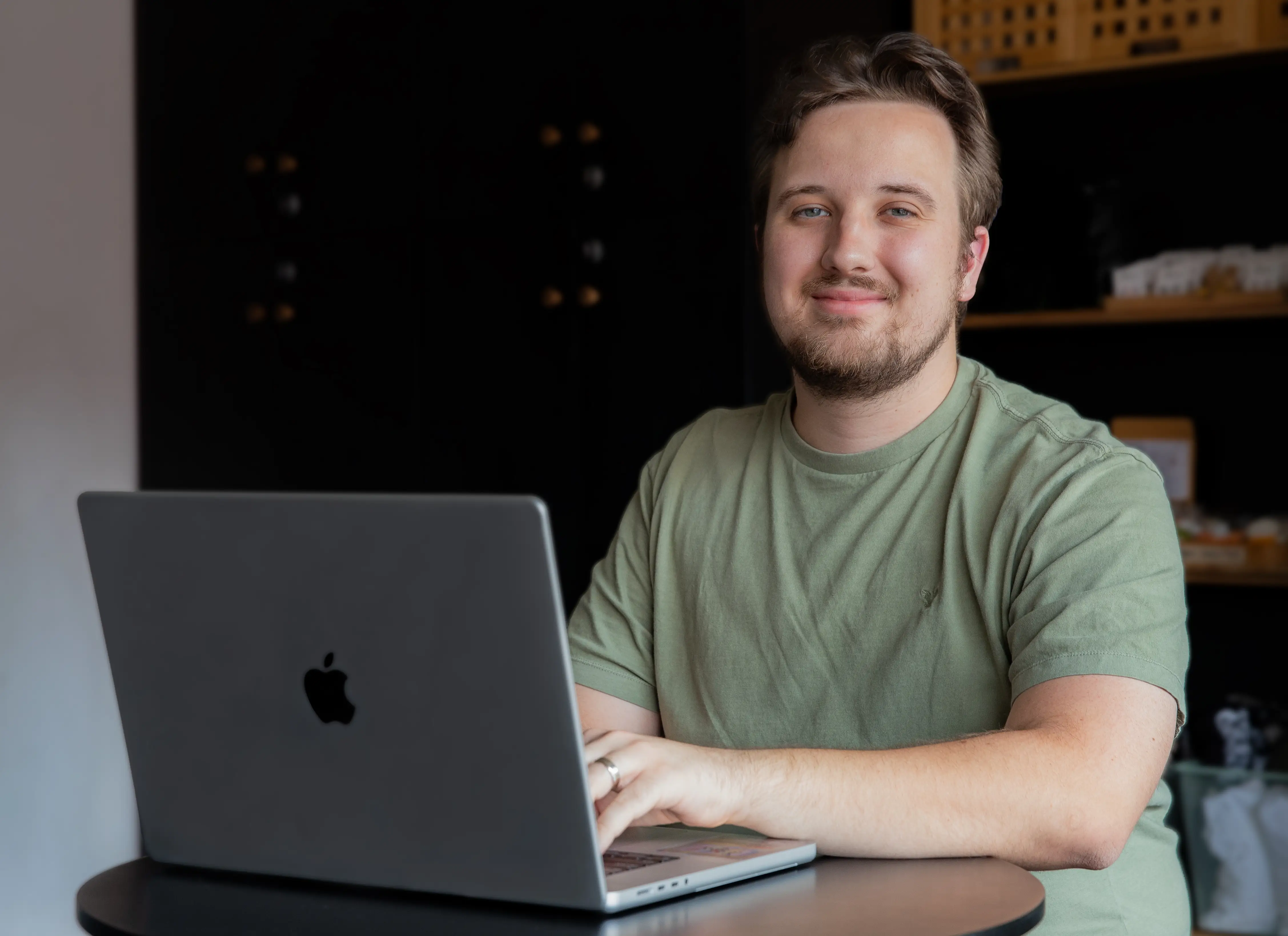 A Caucasian man with light brown hair and a beard smiles as he sits at a table with an open Apple laptop. He's wearing a light green T-shirt.