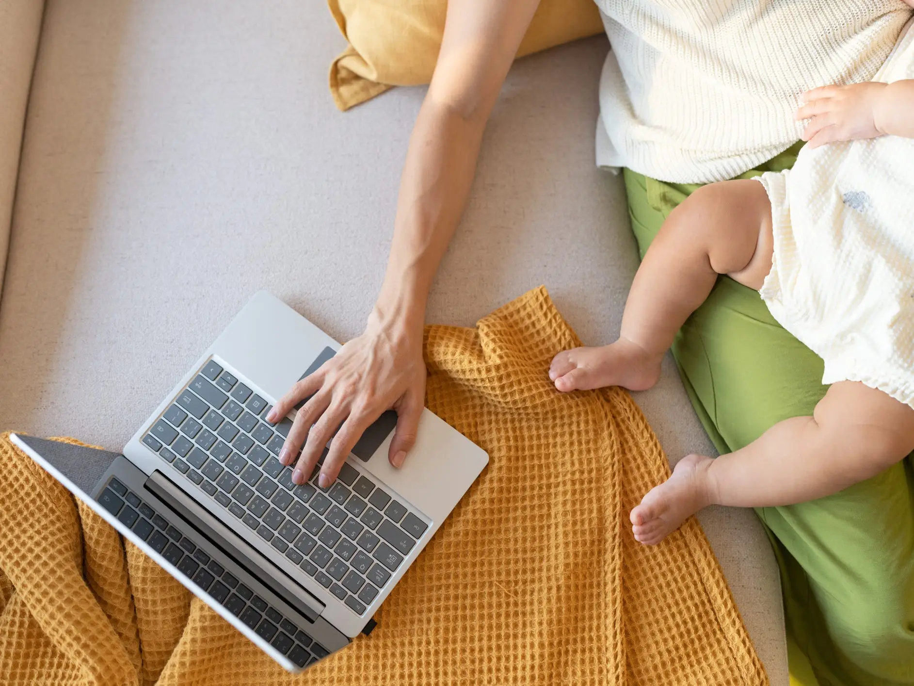 A mother holding a baby on a couch while working on a laptop.