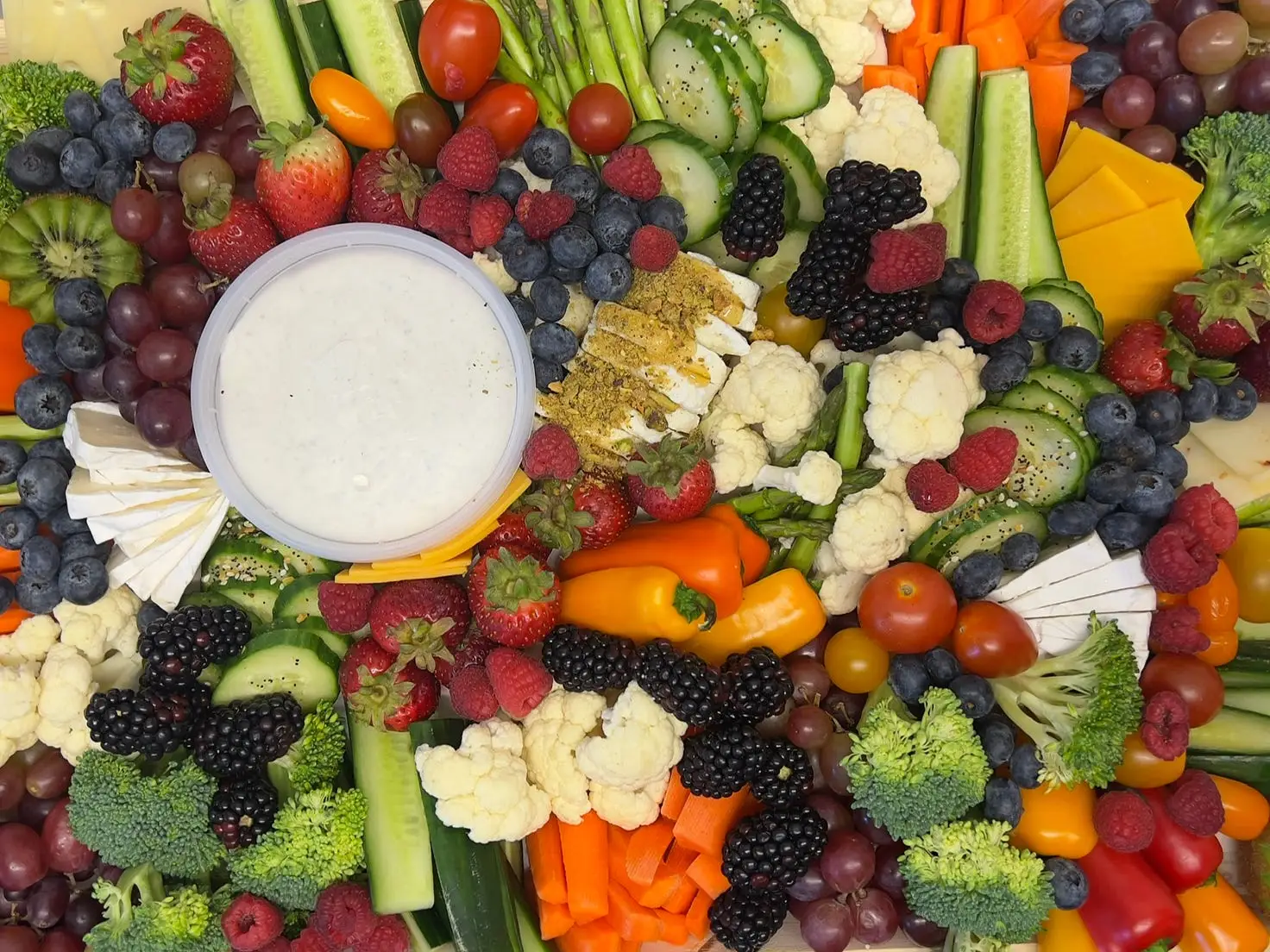 A variety of colorful fruit, vegetables, cheese, and a dip are arranged on a wooden tray.