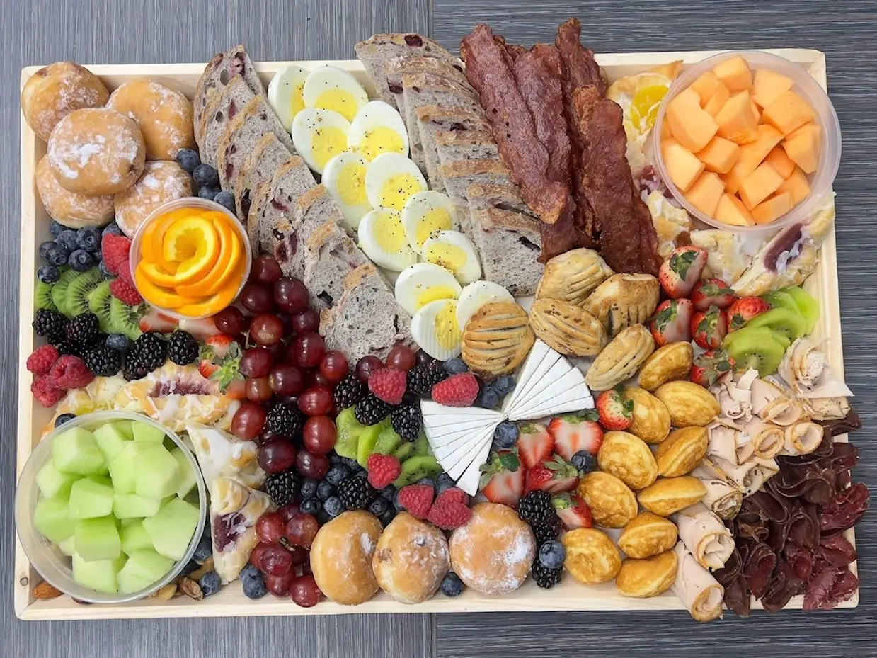 A wooden board filled with fruit, pastries, eggs, bacon, meats, cheeses, and bread sits on a table.
