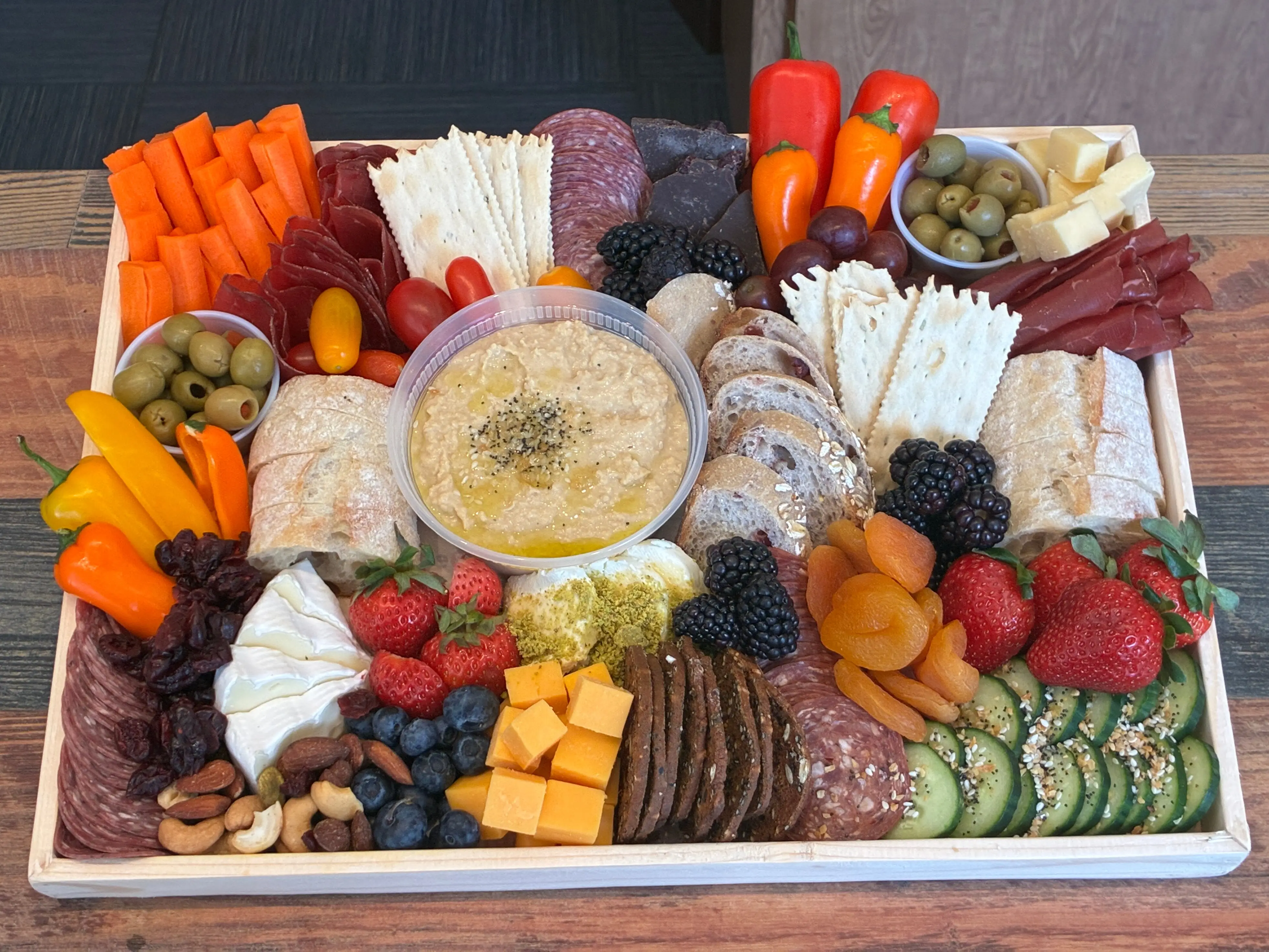 A wooden tray holds a colorful charcuterie spread with meats, cheeses, crackers, fruit, vegetables, olives, bread, and dip.