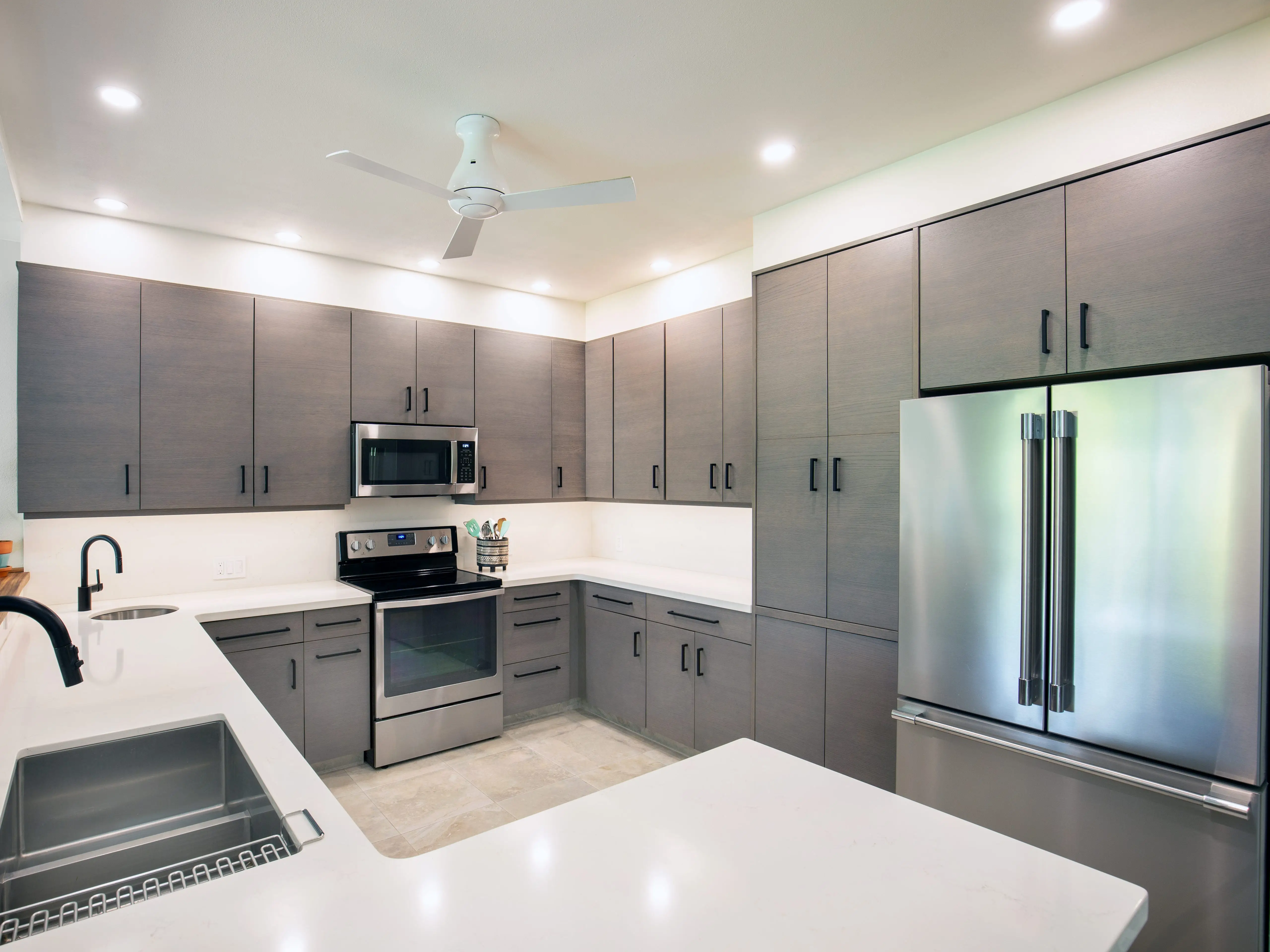Modern kitchen with gray cabinets, stainless steel appliances, white countertops, sinks, black faucets, and recessed ceiling lights.