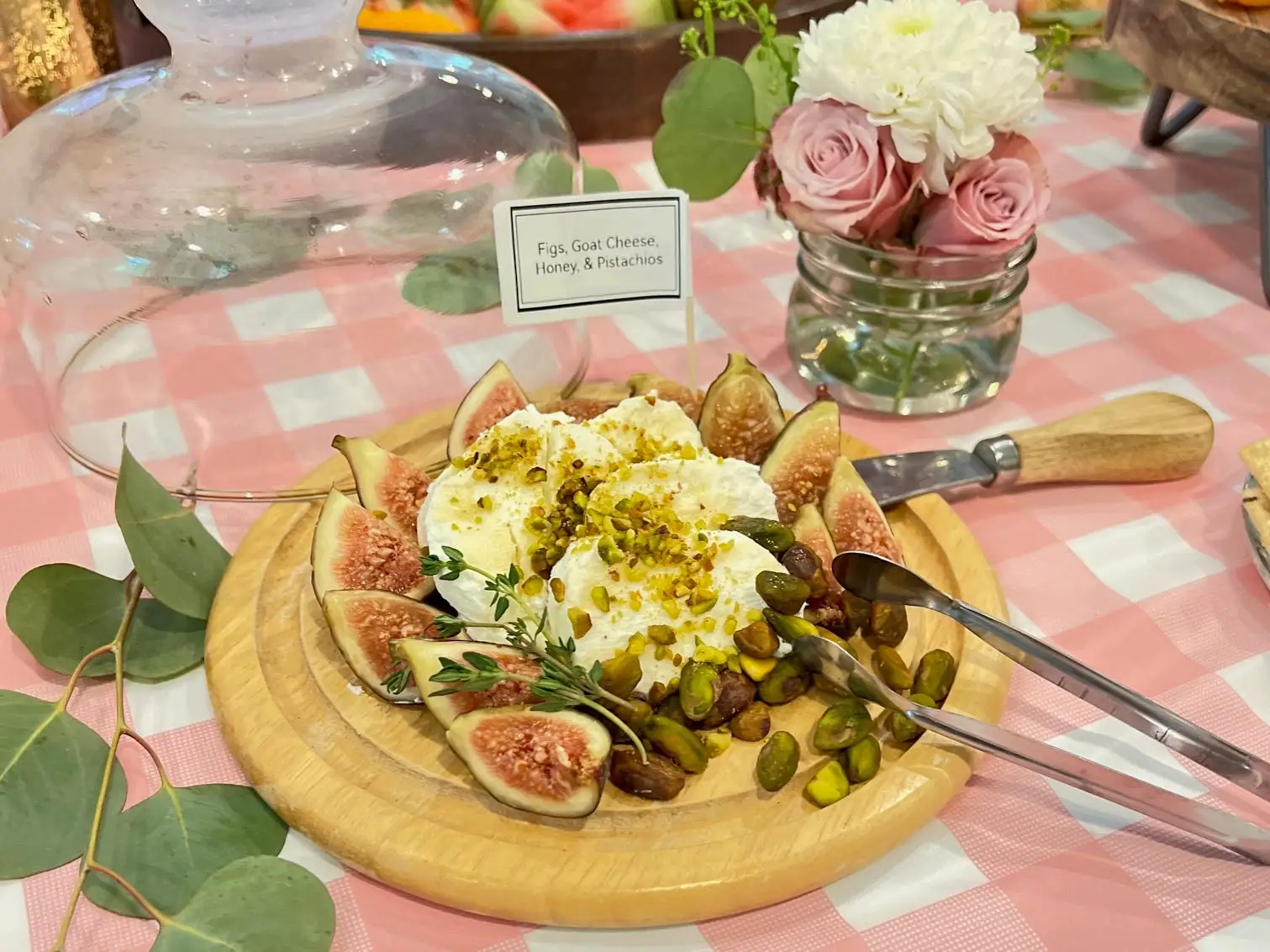 A table with a pink gingham tablecloth holds a cheese plate piled with figs, goat cheese, honey, and pistachios.