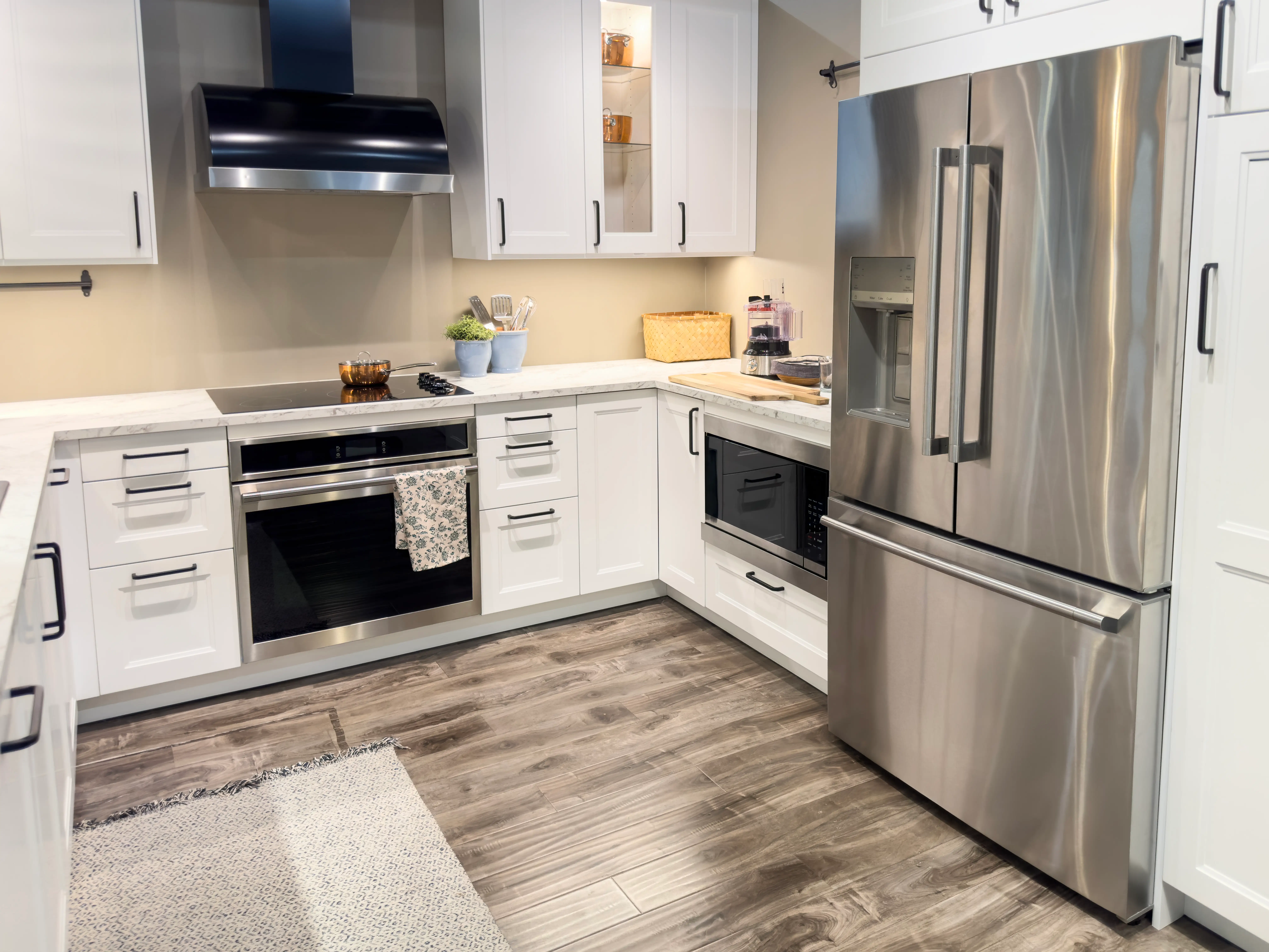 Modern white kitchen with stainless steel refrigerator, range, microwave, hood, shaker cabinets, and wood-look flooring.
