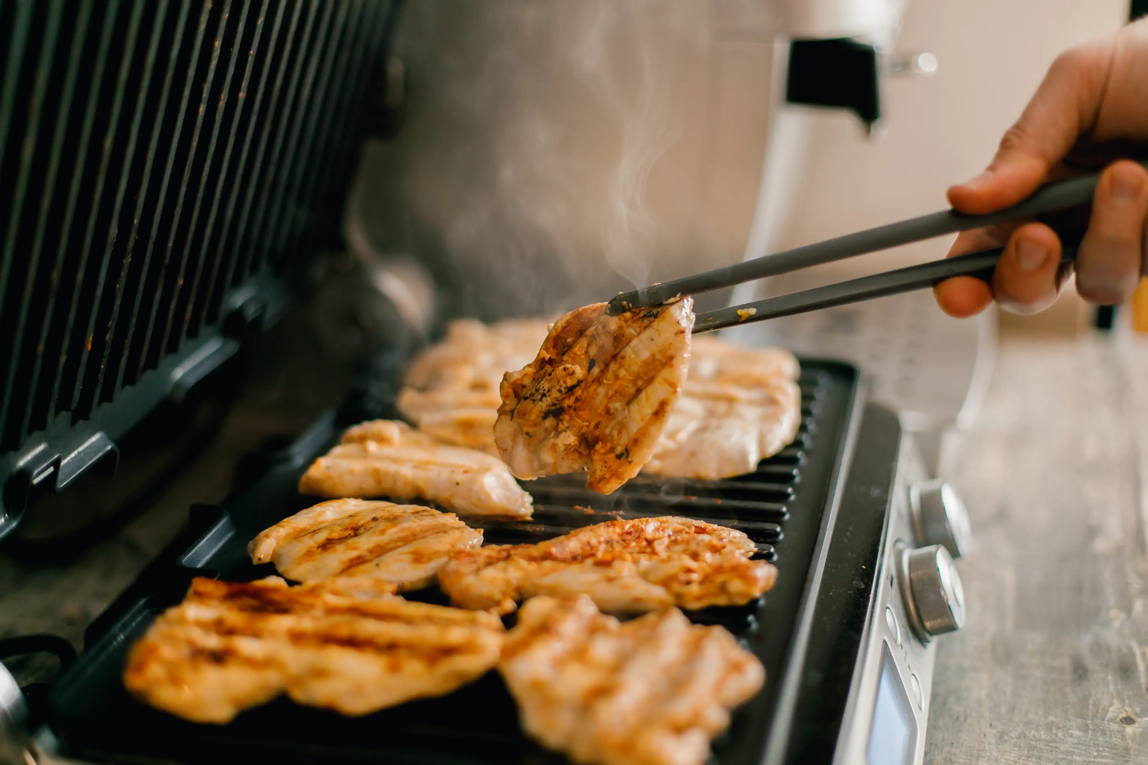 Chicken pieces cook on a steaming indoor grill while a hand lifts one piece with tongs.