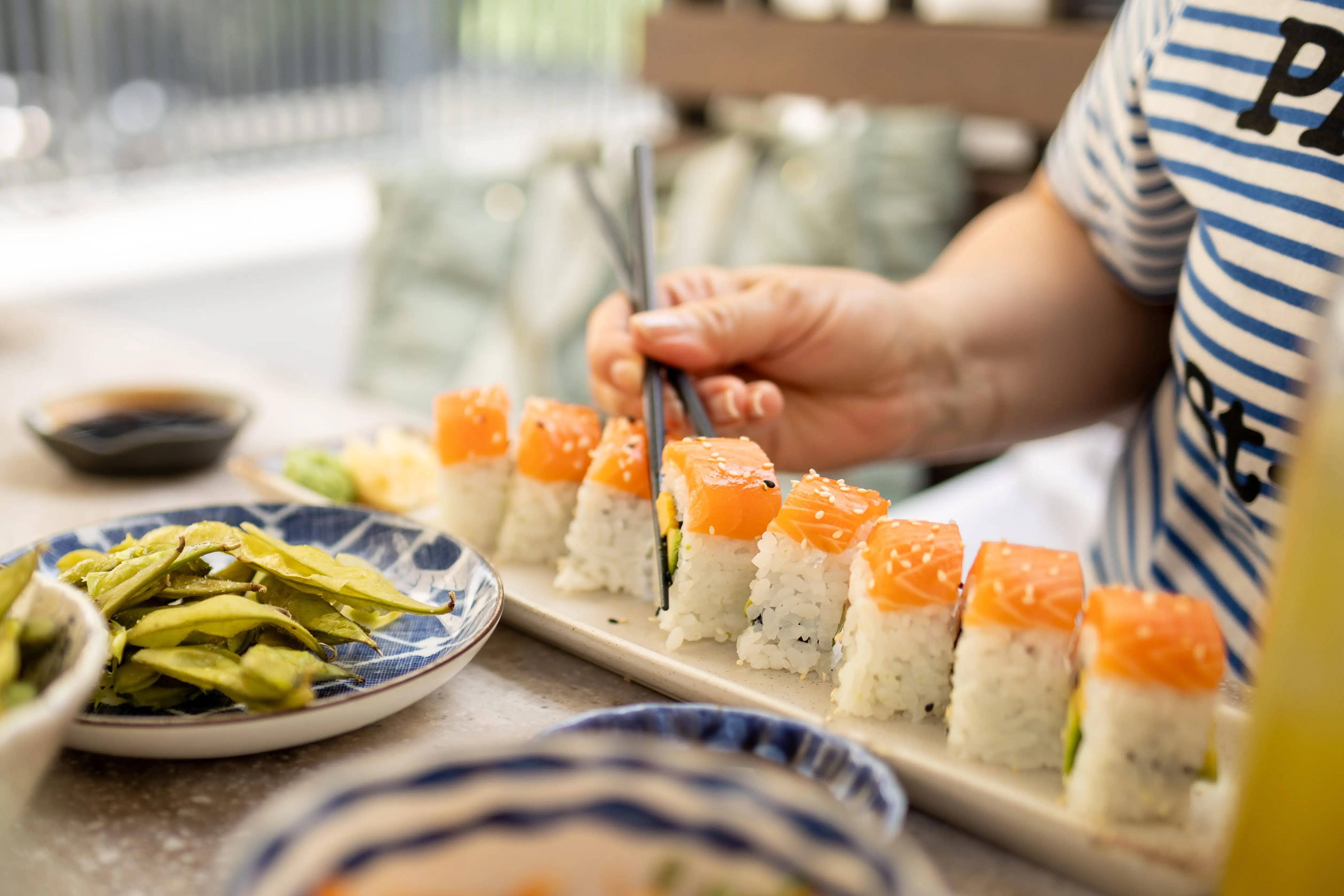 Close-up of a diner using chopsticks to pick up sushi rolls beside edamame and dipping sauces.