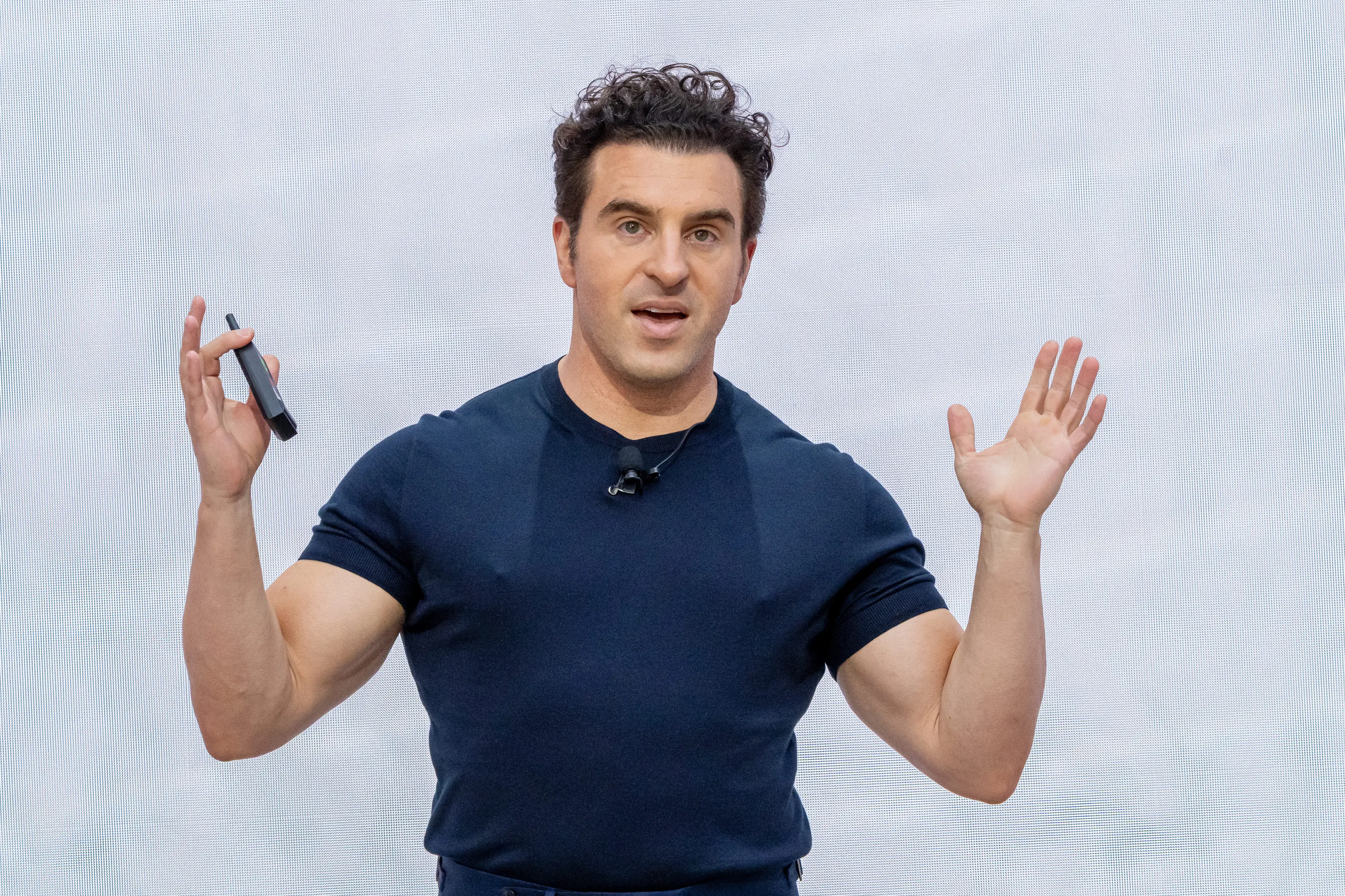 Presenter in a dark shirt holds a clicker with both hands raised against a light stage backdrop.
