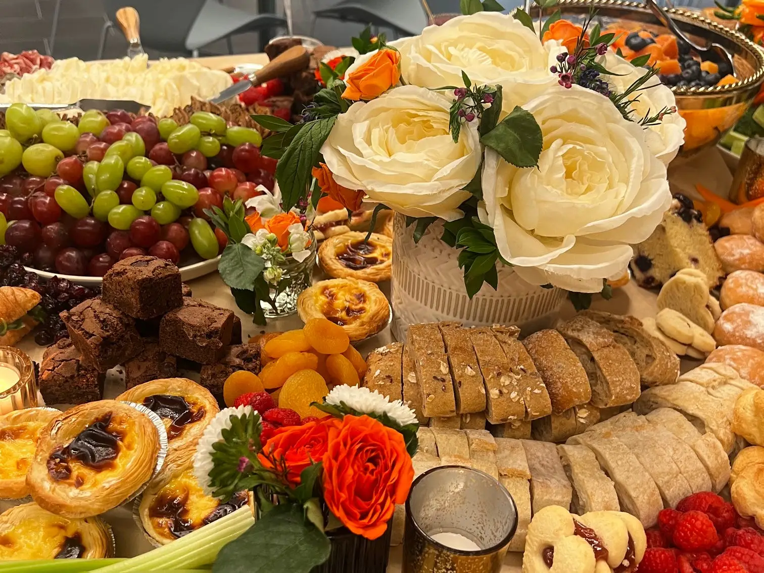 A spread of pastries, fruit, bread, cheese, and flowers is arranged in an event room.