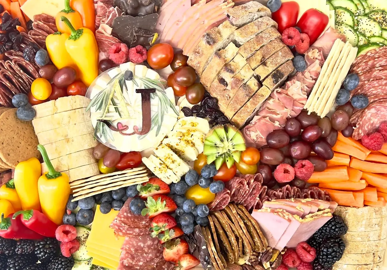 A charcuterie board with a variety of sliced meats, cheeses, crackers, bread, fruit, vegetables, and a decorated cheese wheel sits on a table.