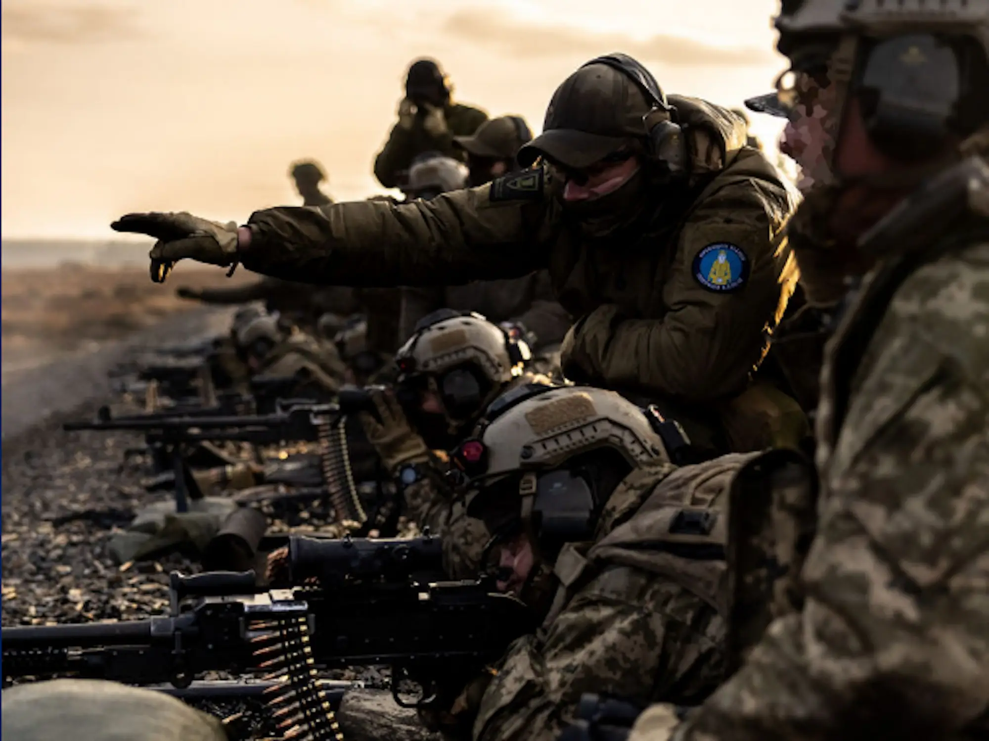 Camouflaged military personnel train with belt-fed machine guns while an instructor points across a rugged outdoor range.
