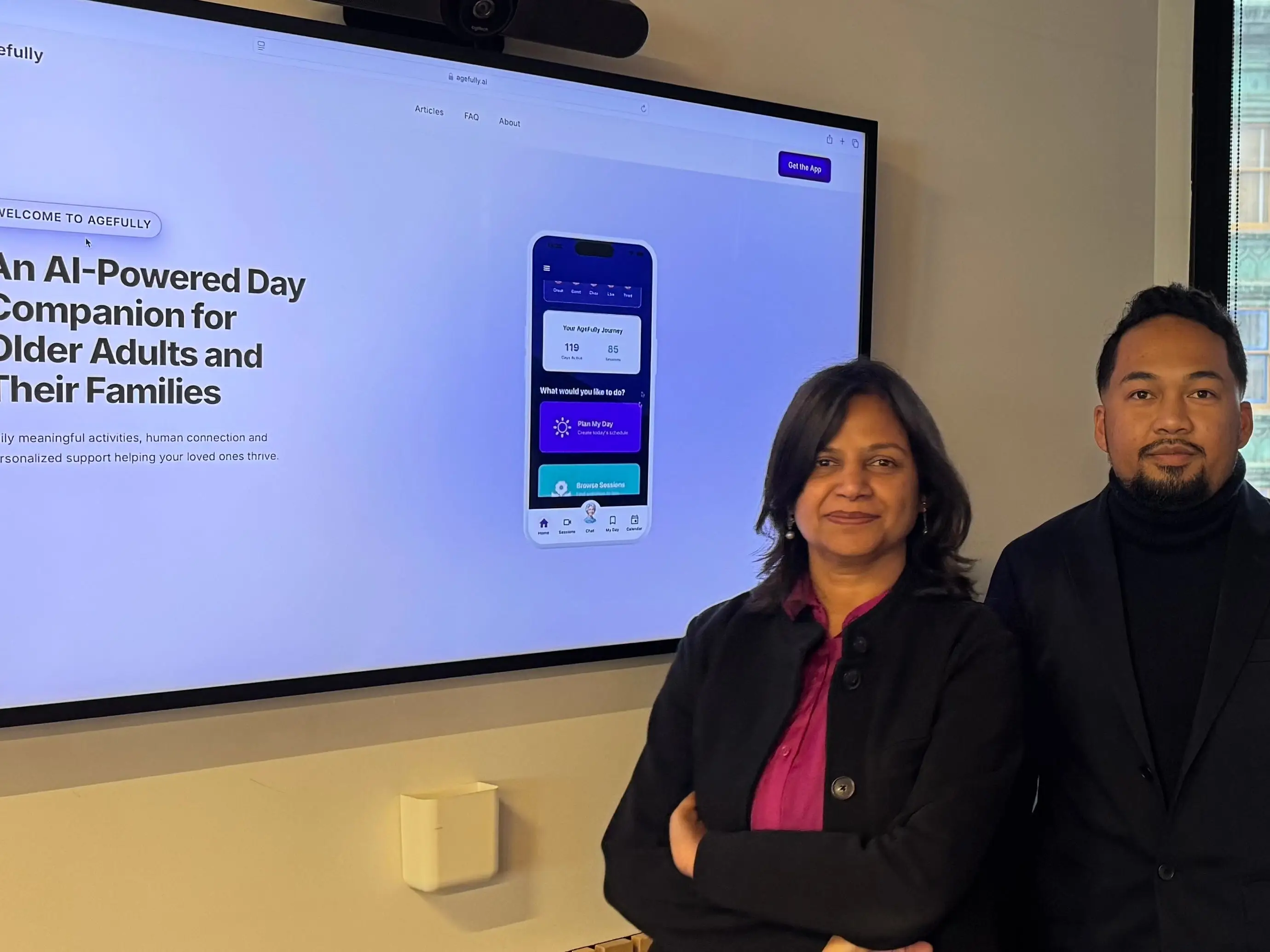 Two people stand beside a large screen displaying the Agefully app, which promotes an AI-powered day companion for older adults and their families.