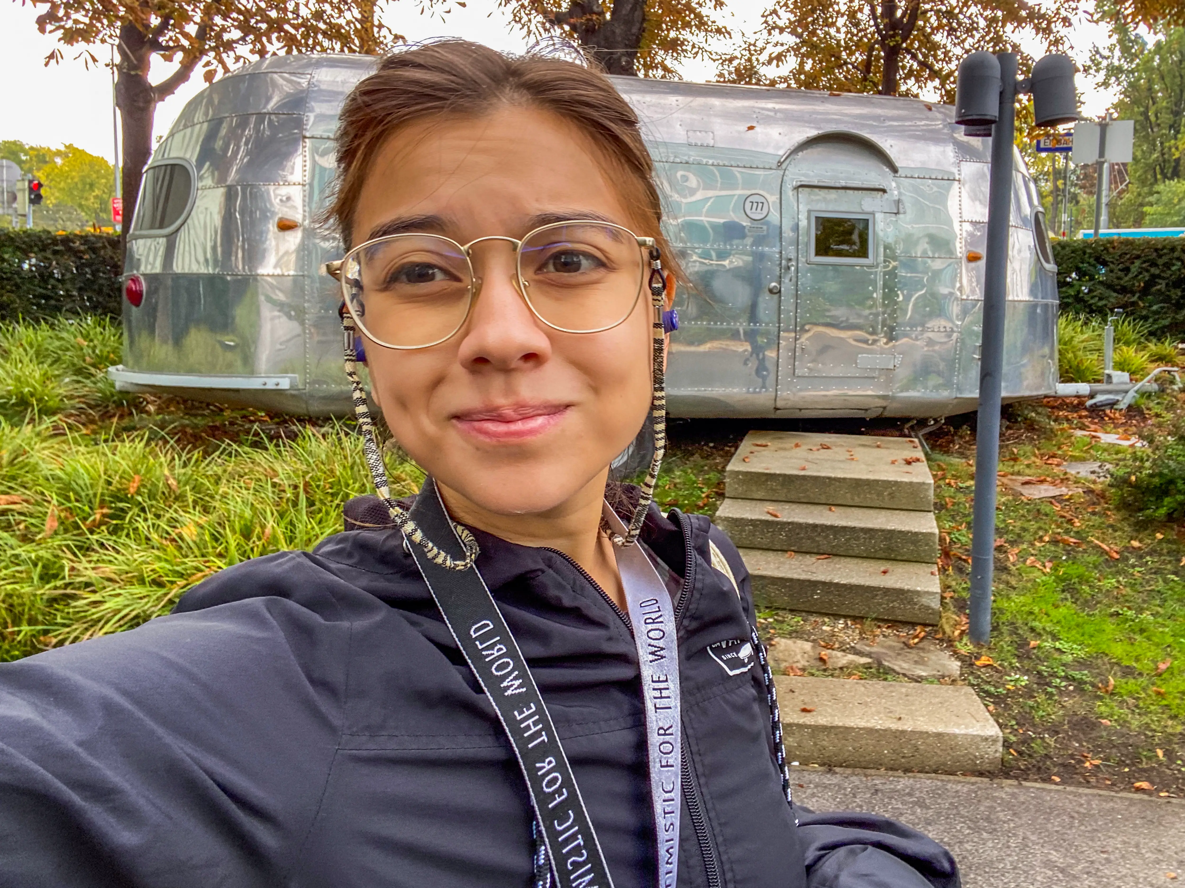 Person taking a selfie in front of a polished aluminum travel trailer on a tree-lined sidewalk.