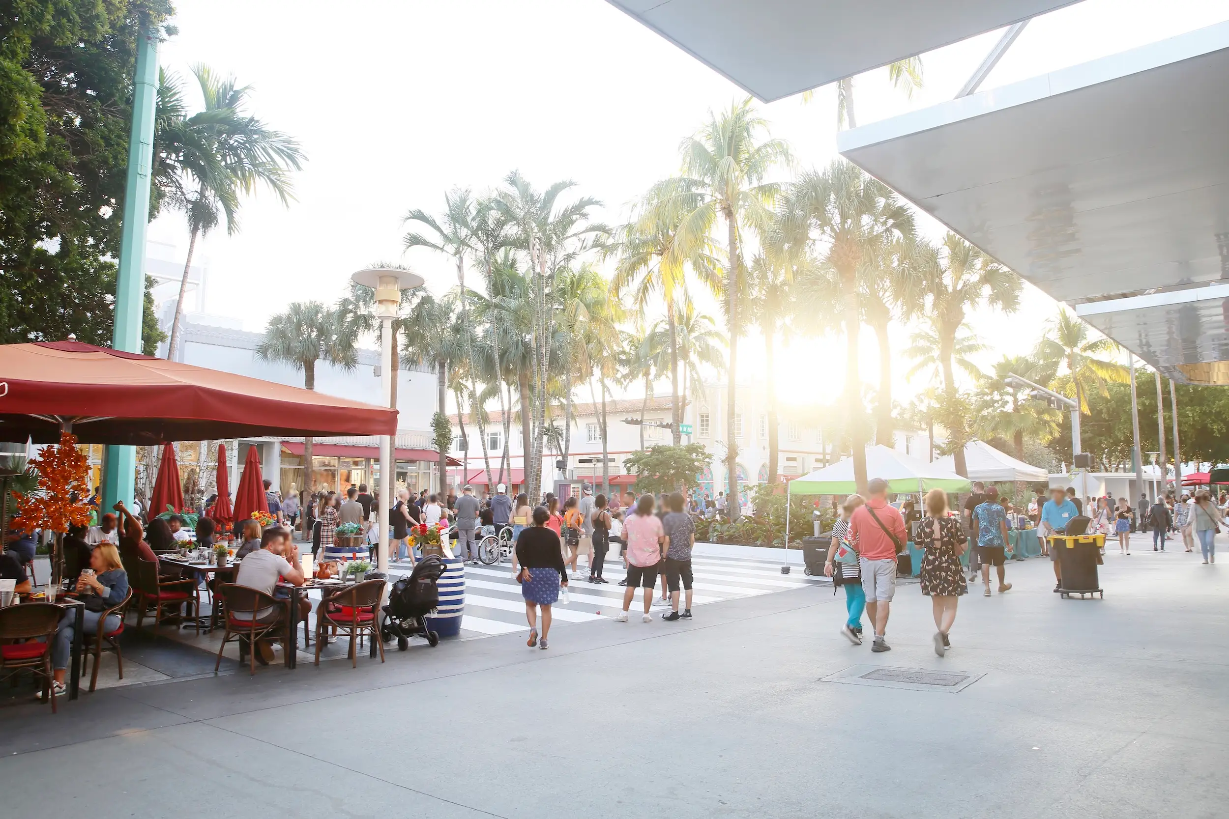 People walk past outdoor café seating and vendor tents on a palm-lined pedestrian street at sunset.