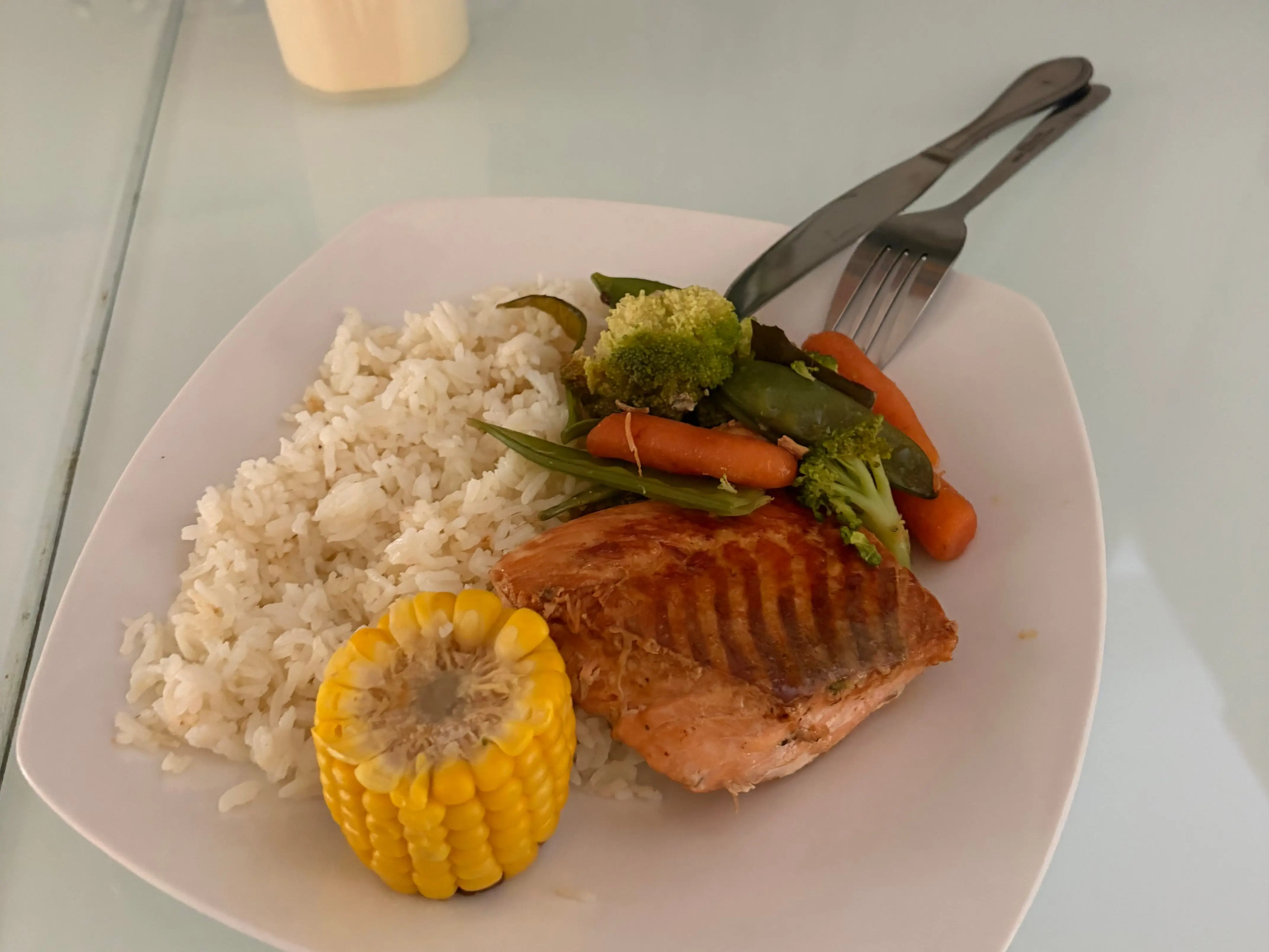 A plate of rice, grilled salmon, corn, and mixed vegetables sits on a glass dining table.