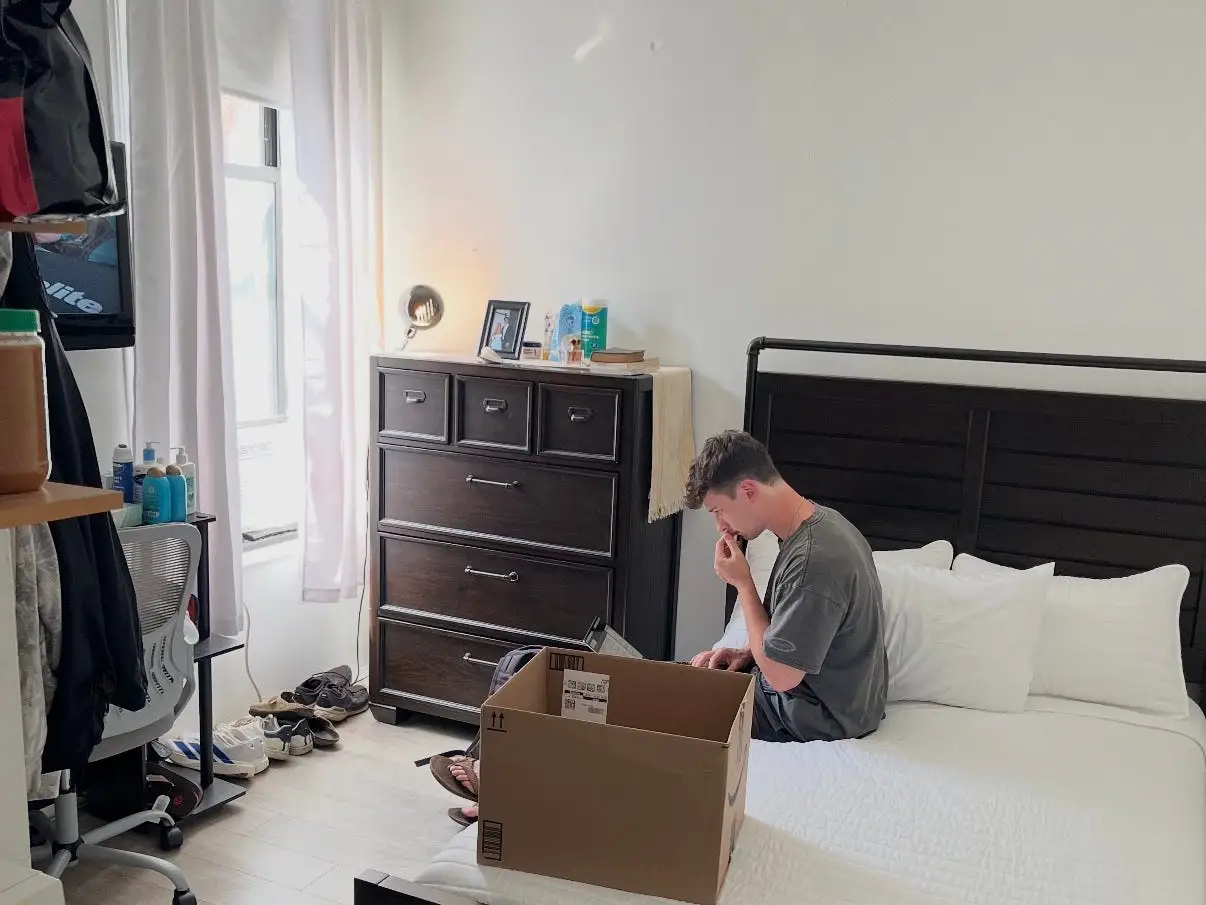 Person sits on a bed beside an open cardboard box in a bedroom with a suitcase and dresser nearby.