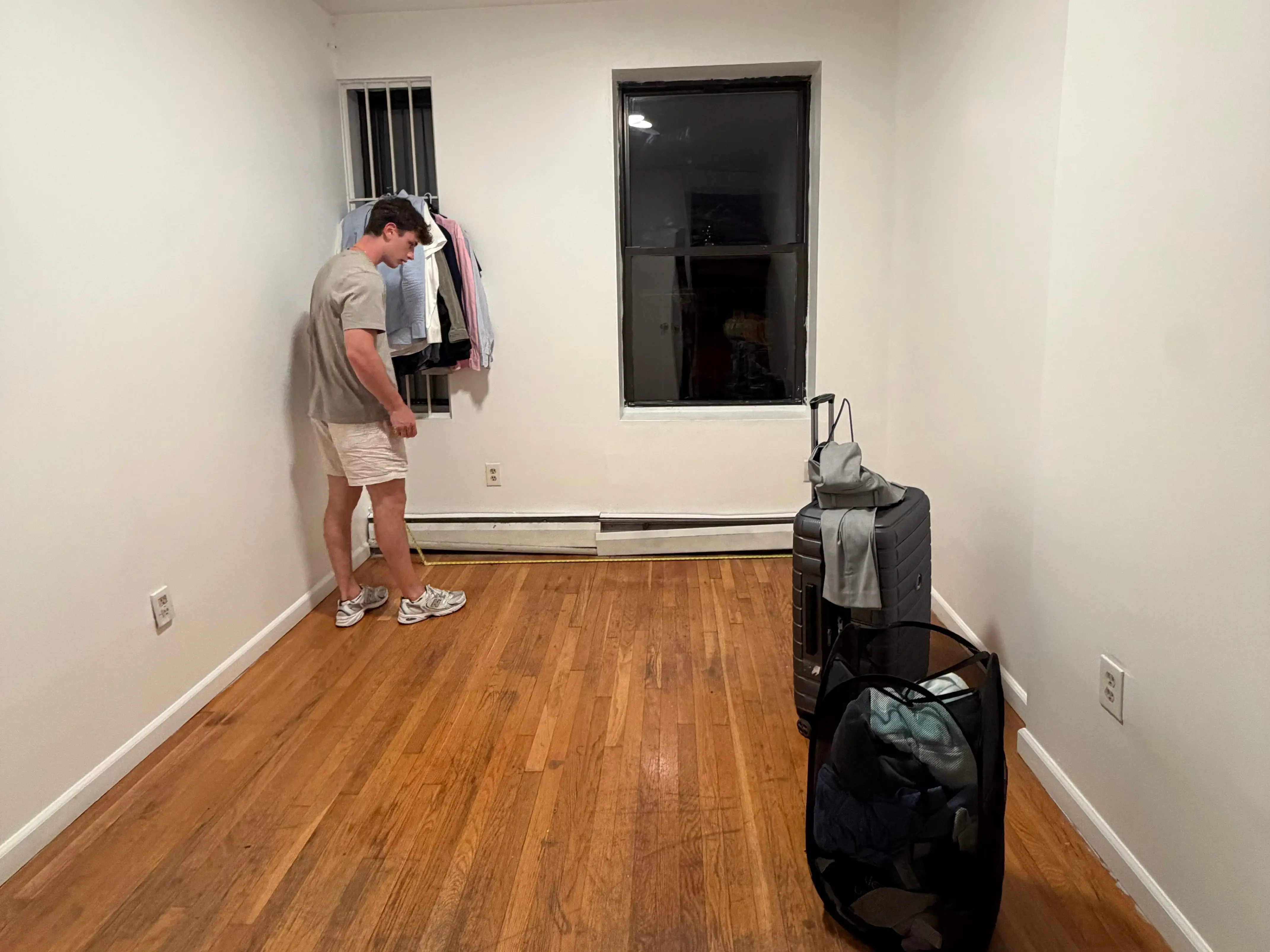 A person stands near hanging clothes in a narrow empty room with wood flooring and luggage.