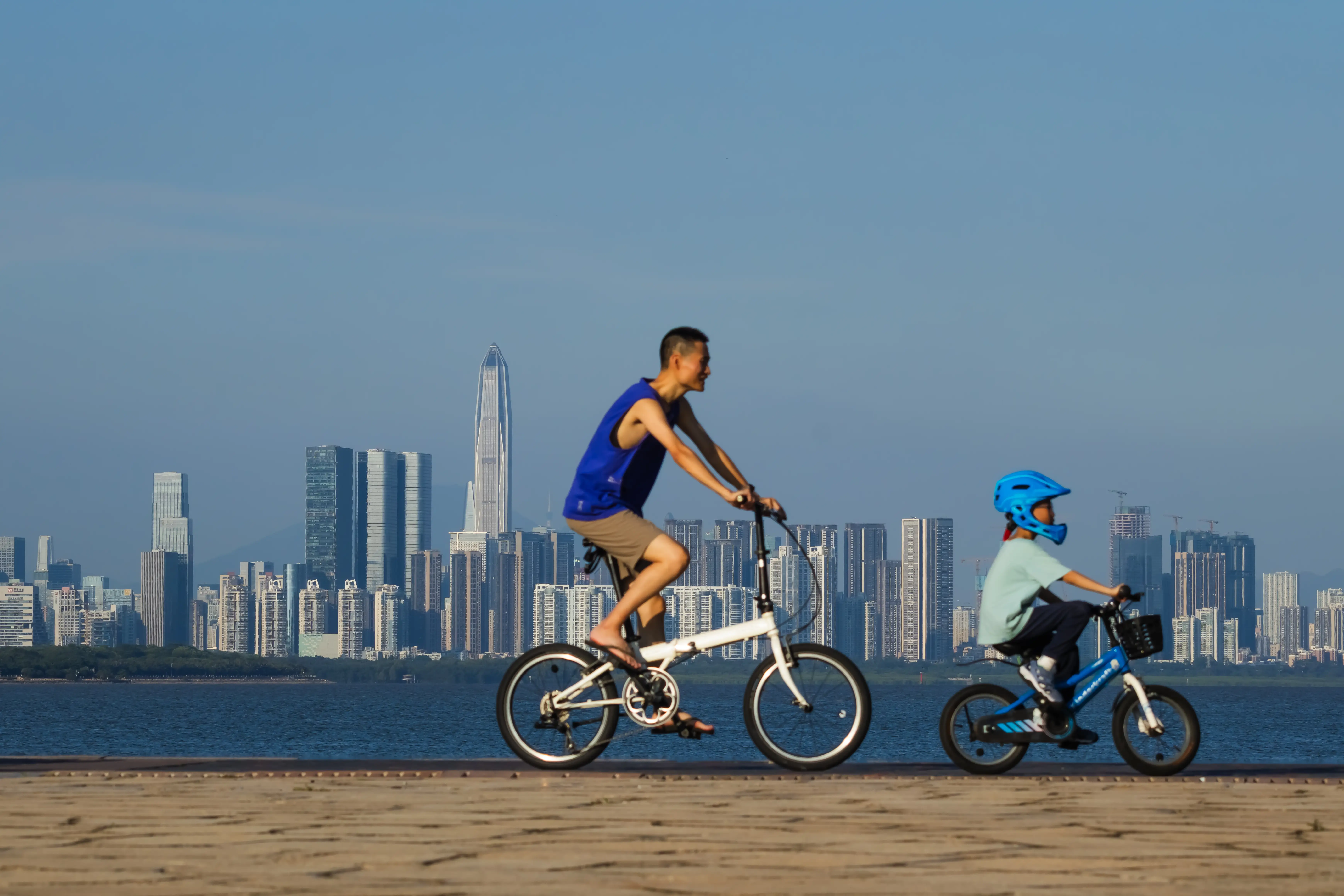 Two cyclists ride along a waterfront promenade with a modern high-rise skyline across the water.