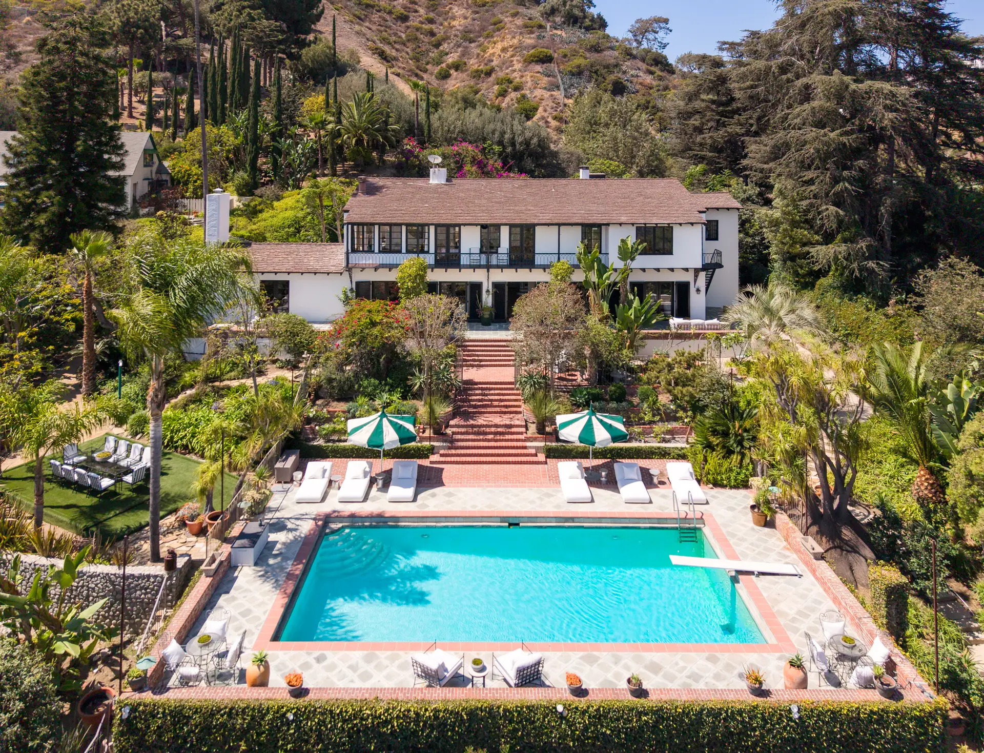 Aerial view of a large hillside villa with a swimming pool, lounge chairs, umbrellas, and lush gardens.