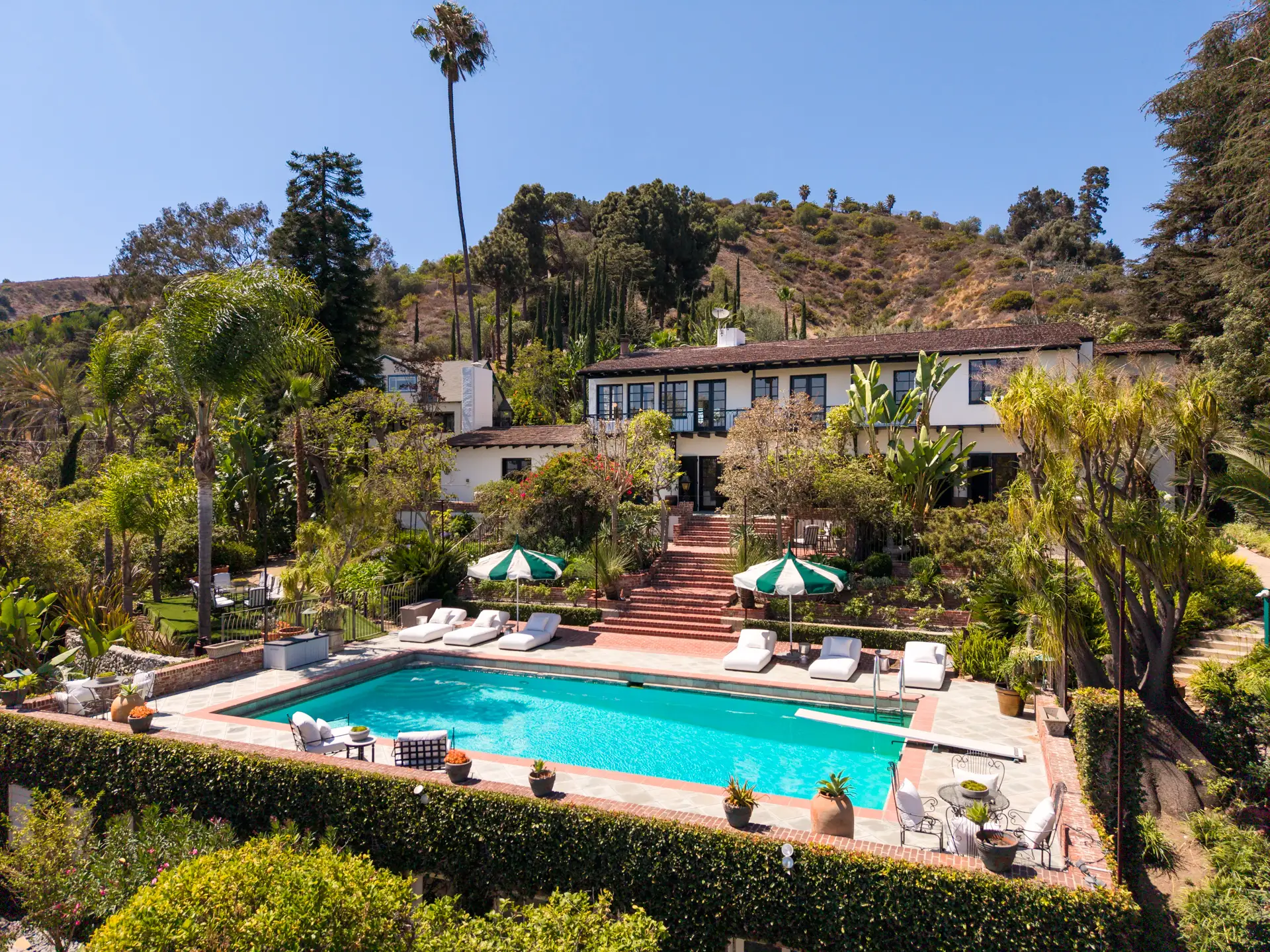 Large hillside estate with a swimming pool, lounge chairs, umbrellas, and lush tropical landscaping under a clear sky.
