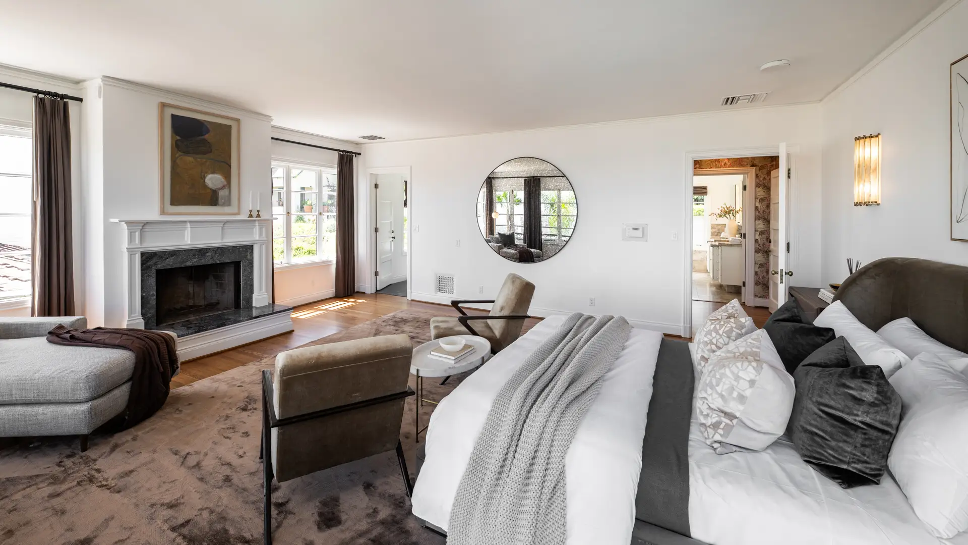 Spacious bedroom suite with a fireplace, large bed, seating area, round wall mirror, and sunlit windows.