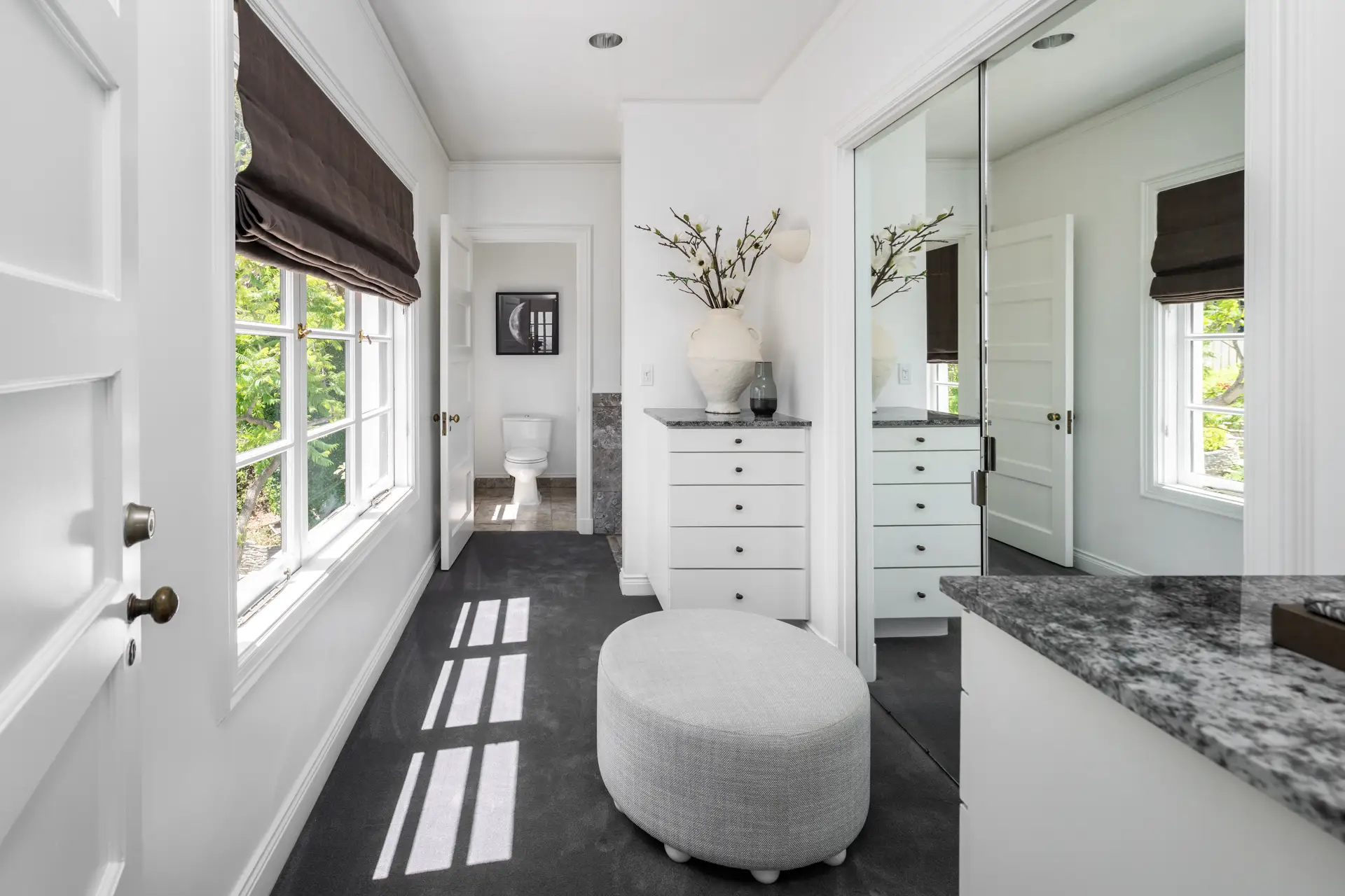 Bright white dressing area with mirrored closet doors, window shades, dresser, ottoman, and adjoining bathroom.