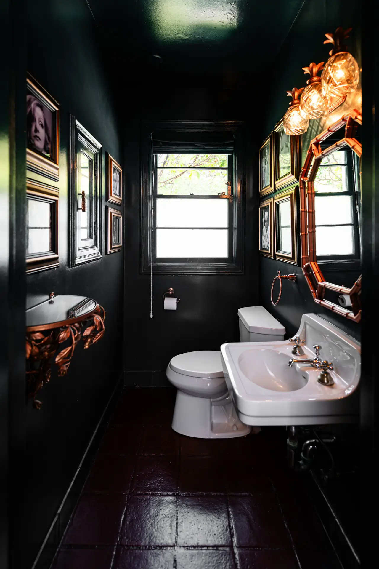 Narrow dark bathroom with white sink and toilet, copper-framed mirror, framed portraits, windows, and warm wall lights.