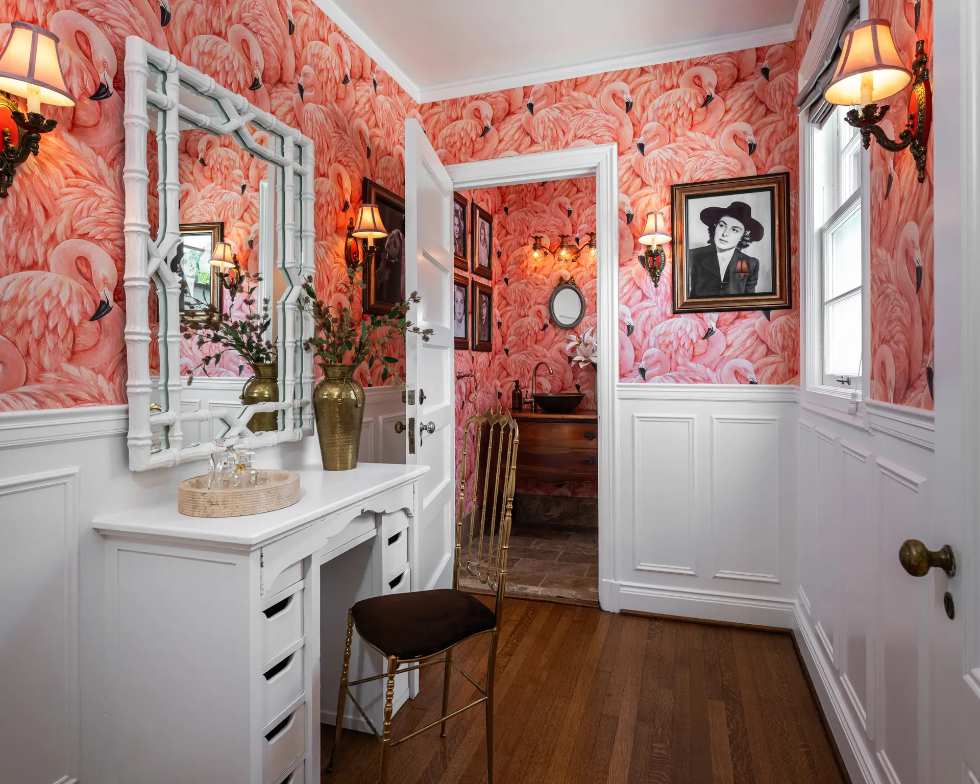 Narrow dressing area with pink flamingo wallpaper, white wainscoting, vanity table, mirror, chair, sconces, and framed portraits.
