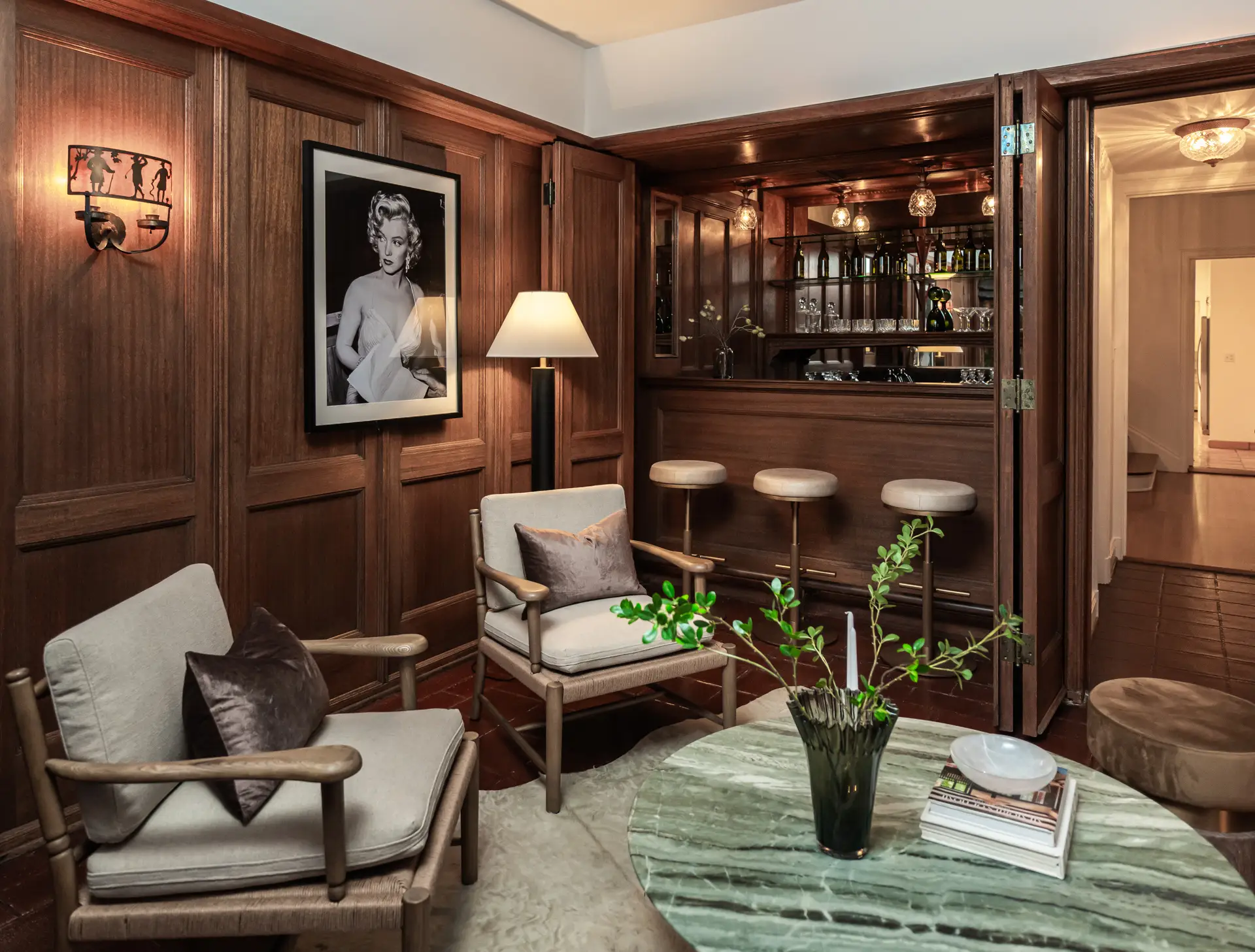 Wood-paneled lounge with chairs, green marble table, home bar, lamps, and a framed Marilyn Monroe portrait.