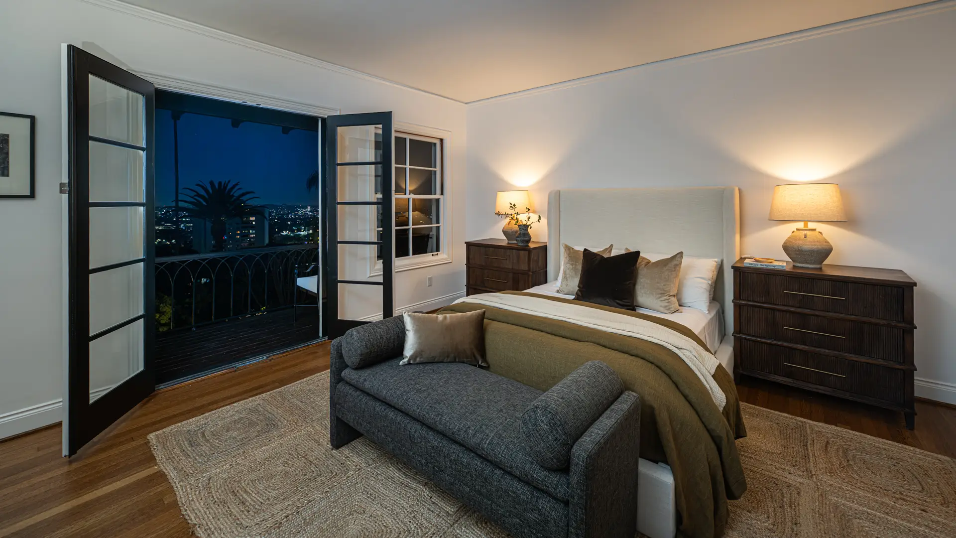 Bedroom with a made bed, bedside lamps, gray bench, and open balcony doors overlooking city lights at night.