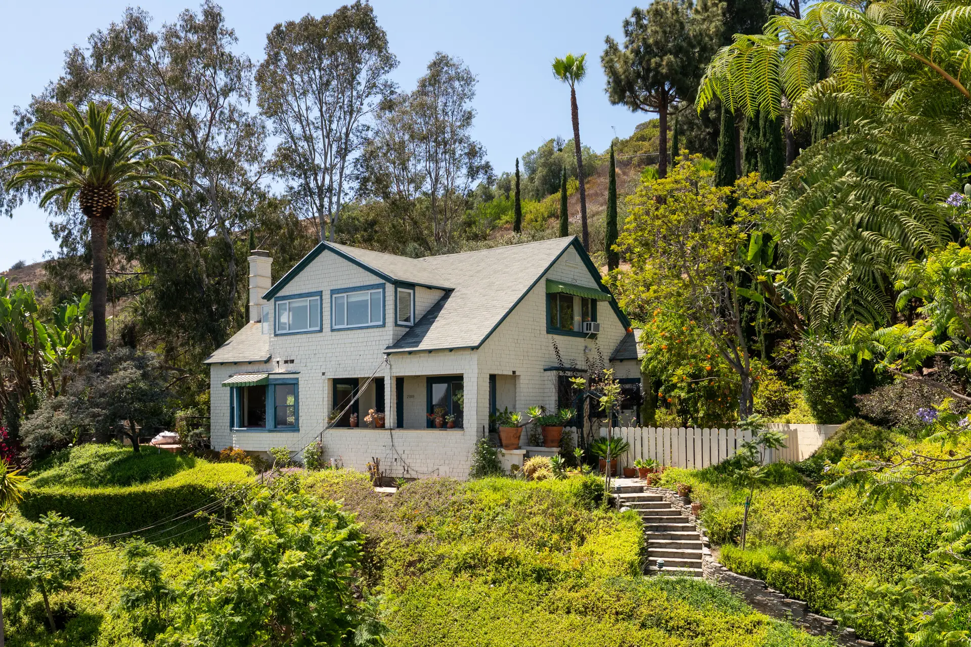Two-story house with green trim sits among dense hillside gardens, palm trees, and outdoor steps.